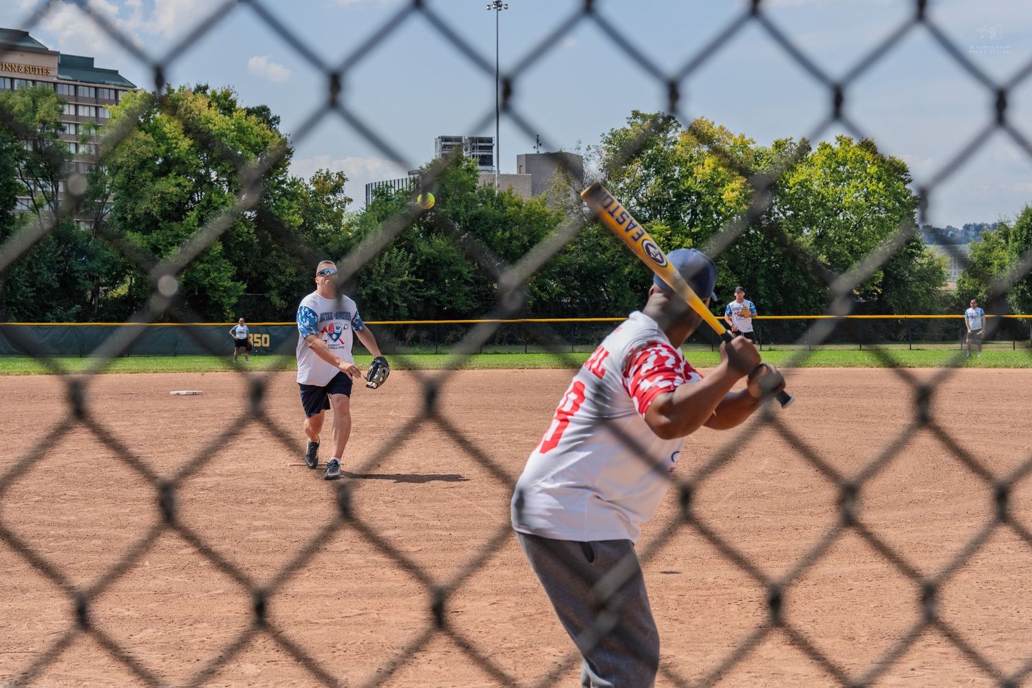Softball batter preparing to swing at a pitch viewed through a chain-link fence during a game.