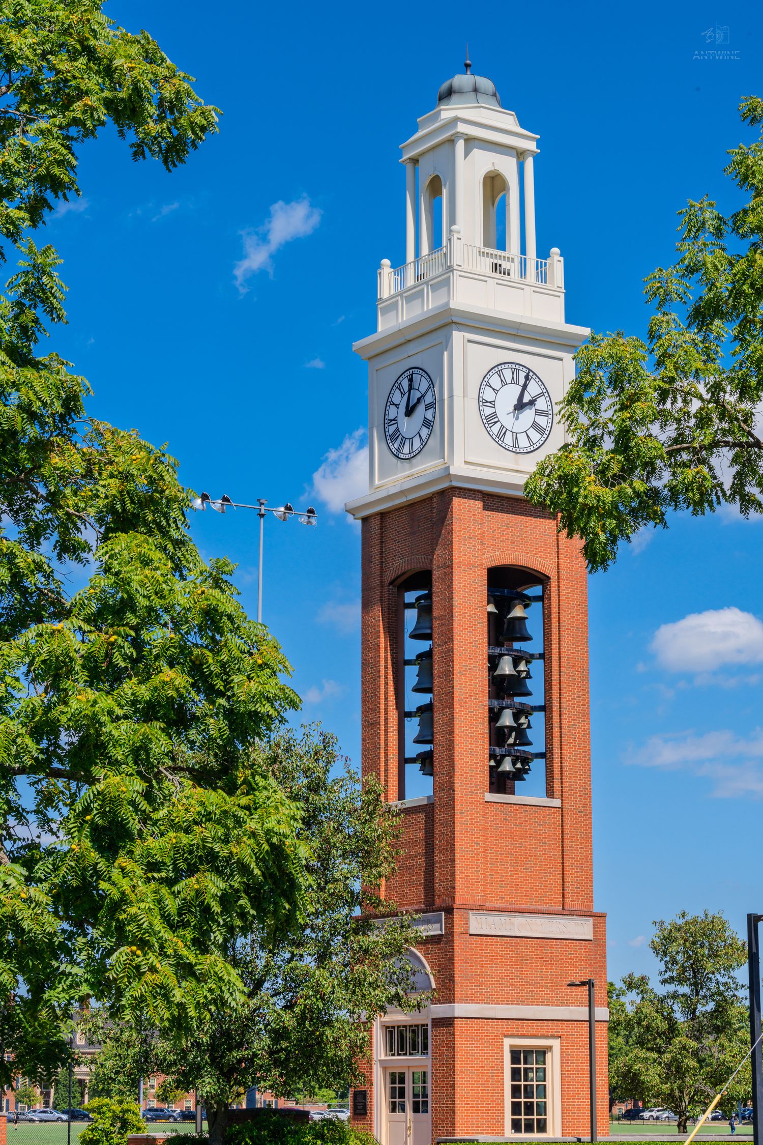 Brick clock tower with white cupola and bells surrounded by green trees against a bright blue sky.