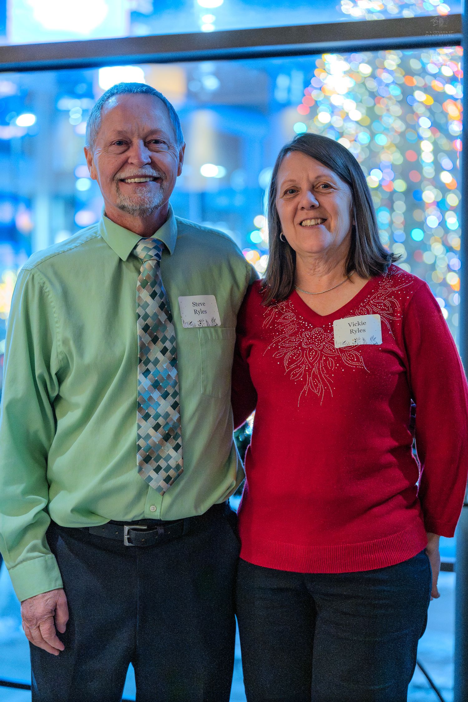 A smiling man in green shirt and tie poses with a woman in red top at a festive holiday event.