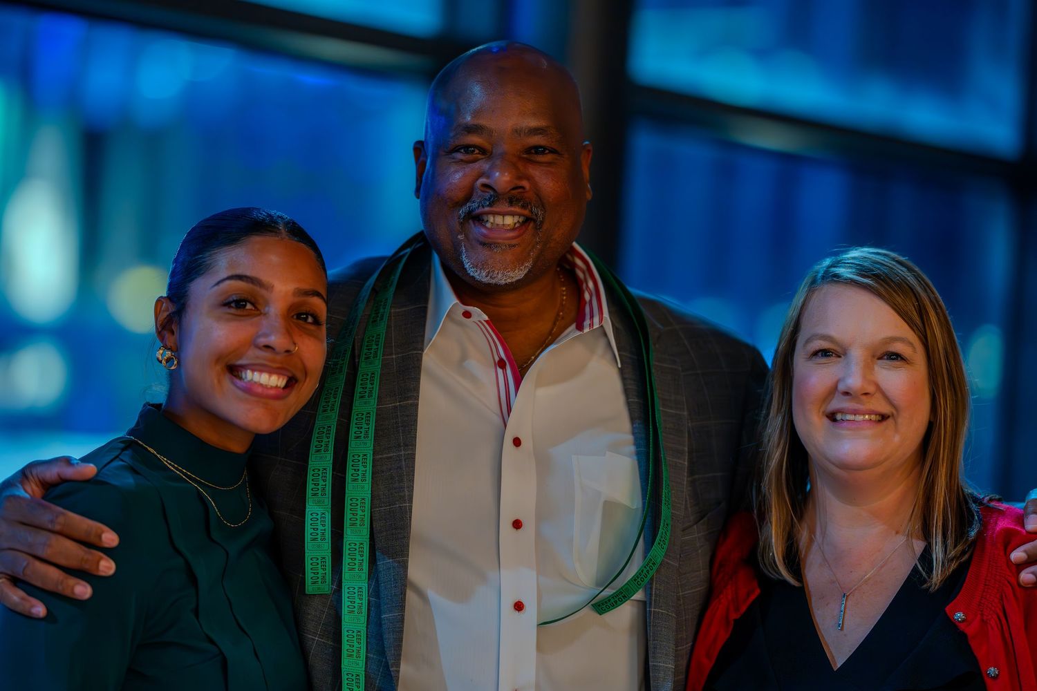 Three smiling people pose together at an indoor event, with a man wearing a green sash flanked by two women.