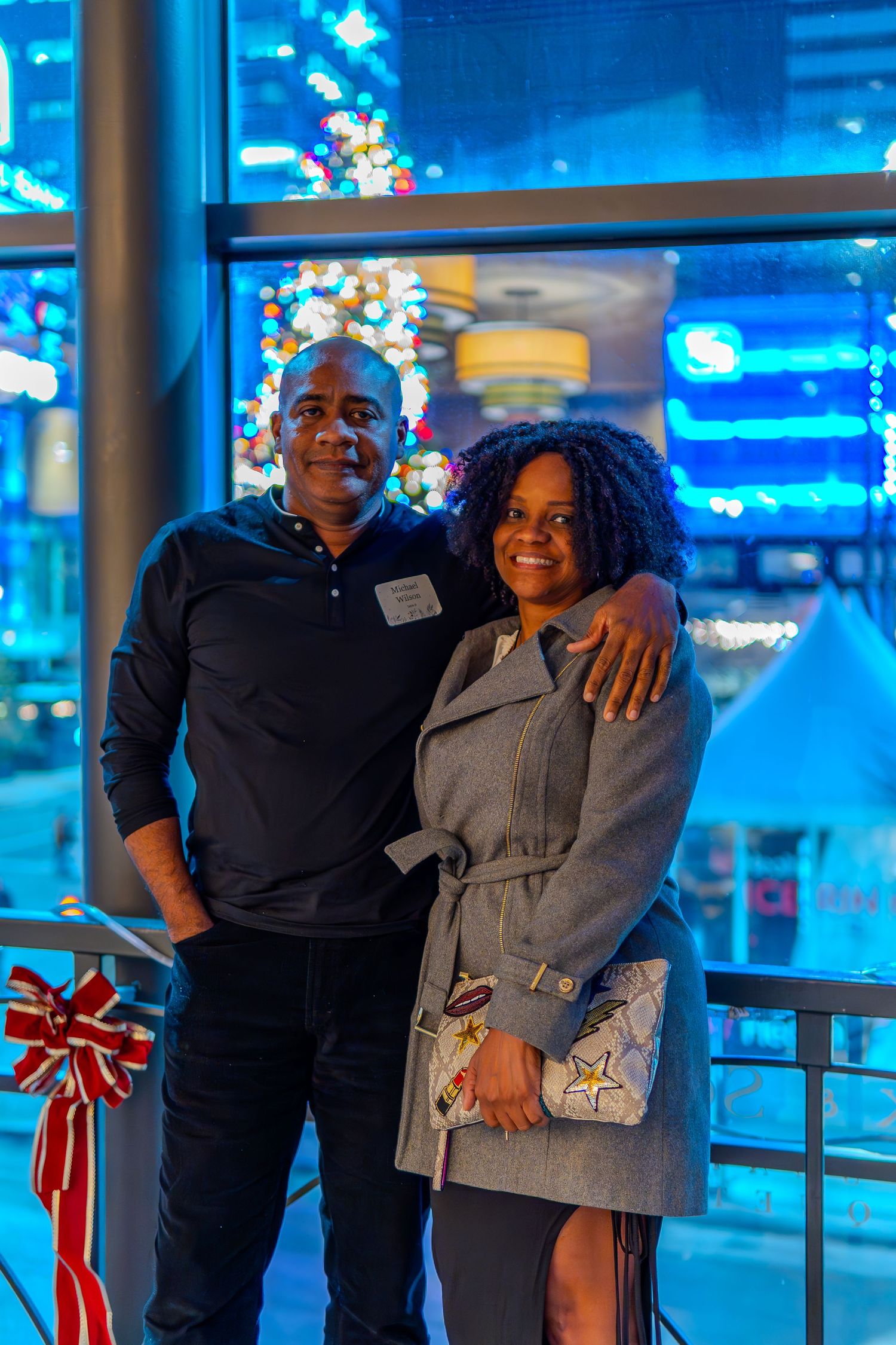 Couple smiling together at a festive holiday event with a lit Christmas tree and city lights in the background.