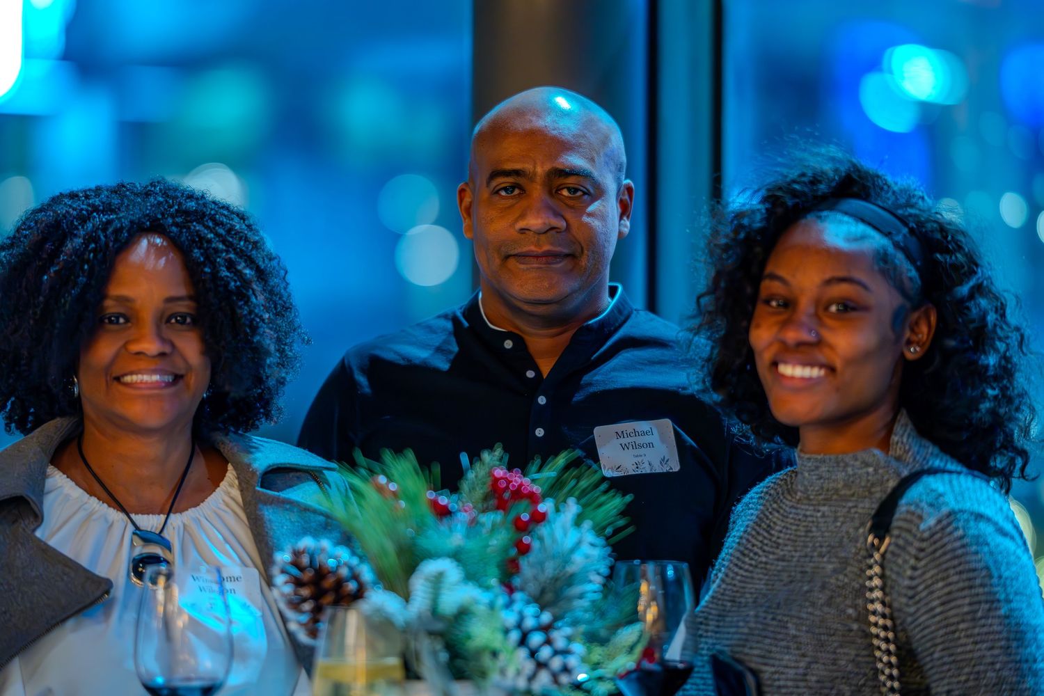 Three people smiling together at an evening event with blue ambient lighting and festive table decor.