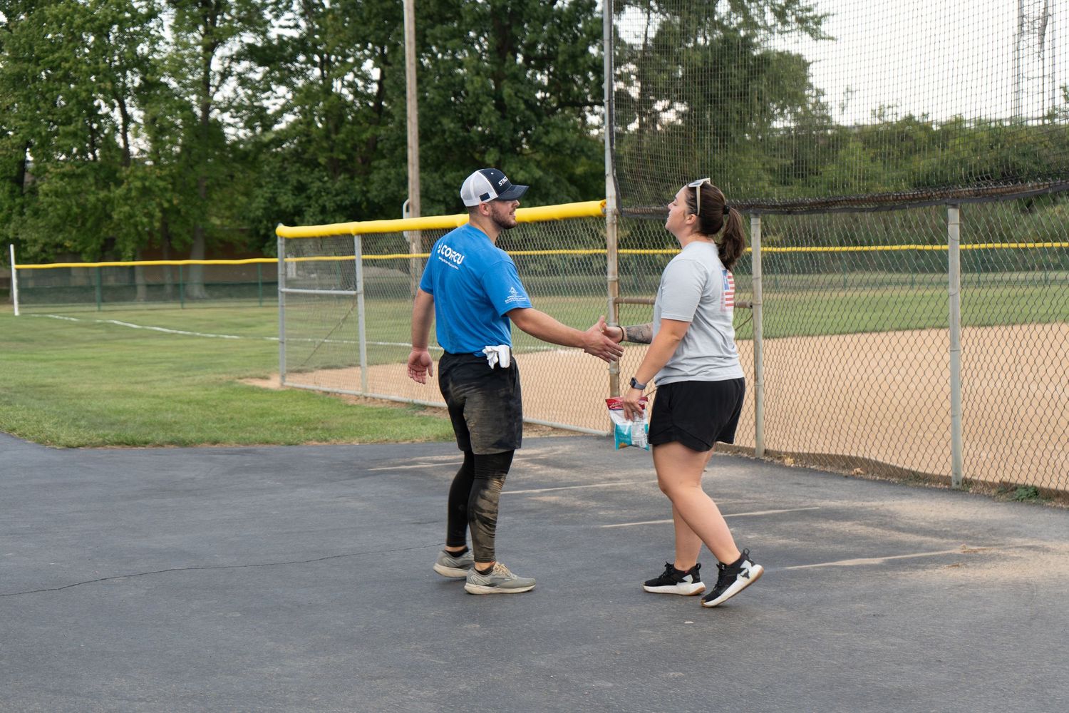 Two people shaking hands on a baseball field blacktop near a chain-link fence on a sunny day.