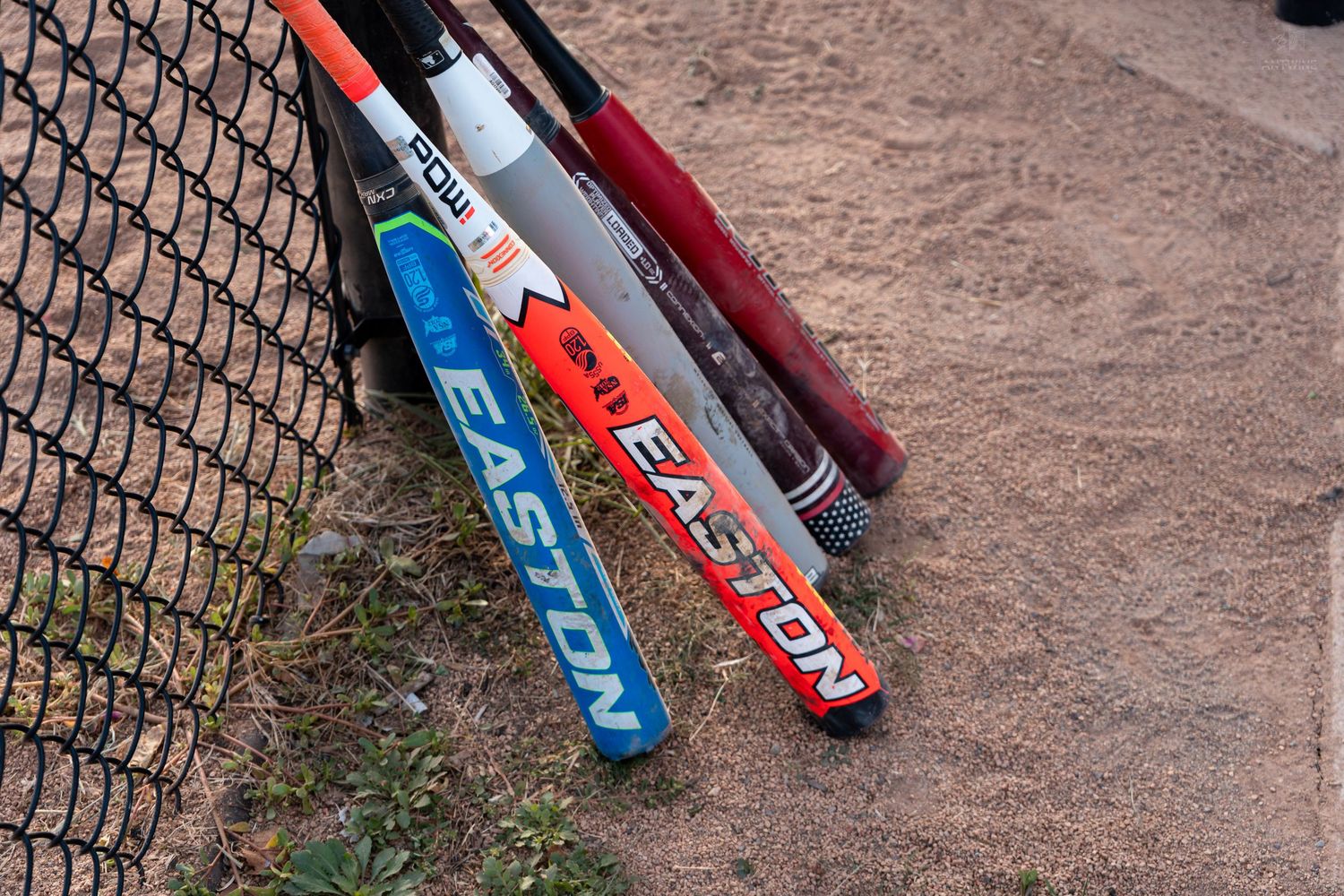 Three Easton softball bats leaning against a chain-link fence on a sandy baseball field.