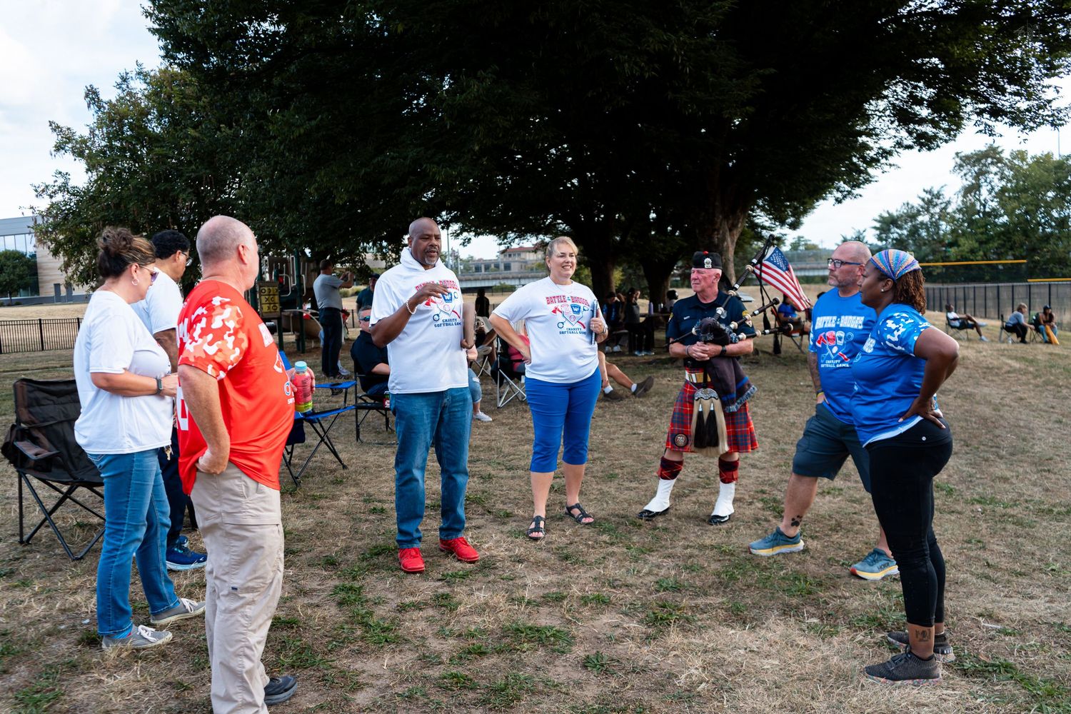 Diverse group of people gather outdoors at a community event near trees and American flag.