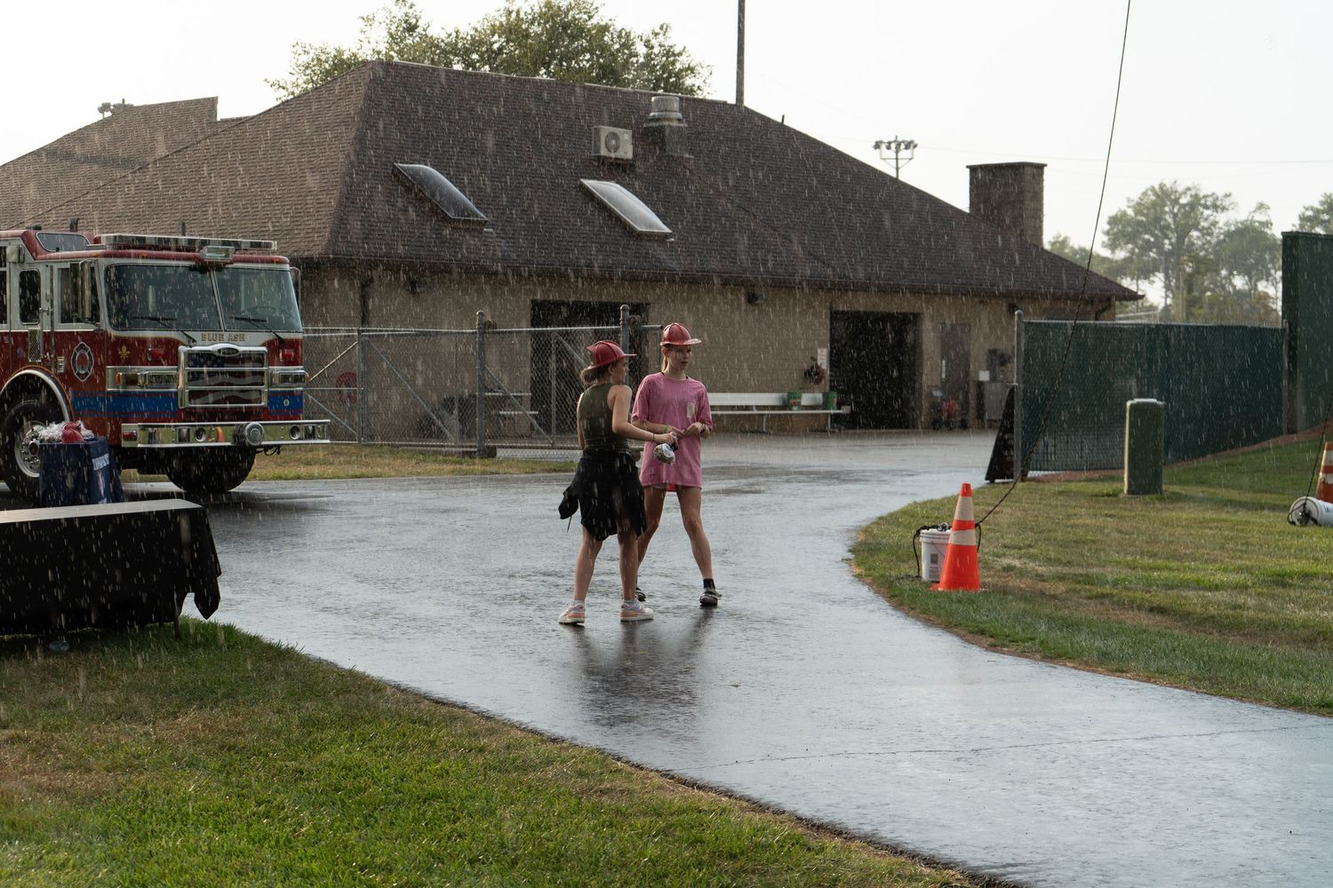 Two women walking on a wet rainy day near a fire station building with a red fire truck visible.