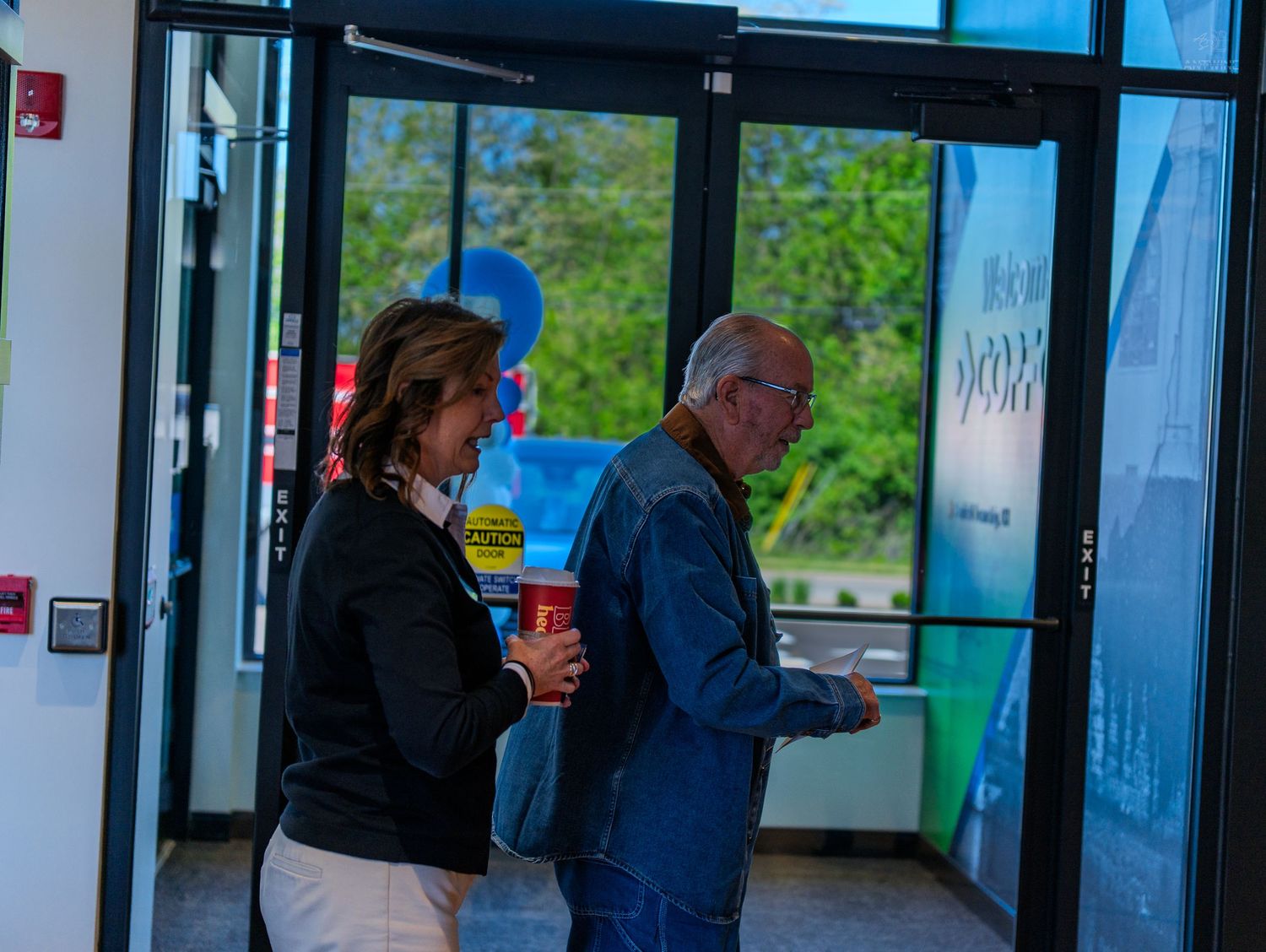 Two people entering a modern building through glass doors at a credit union grand opening event.