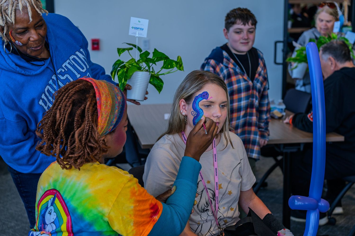 A child gets blue face paint applied at a colorful indoor community event with people gathered around.