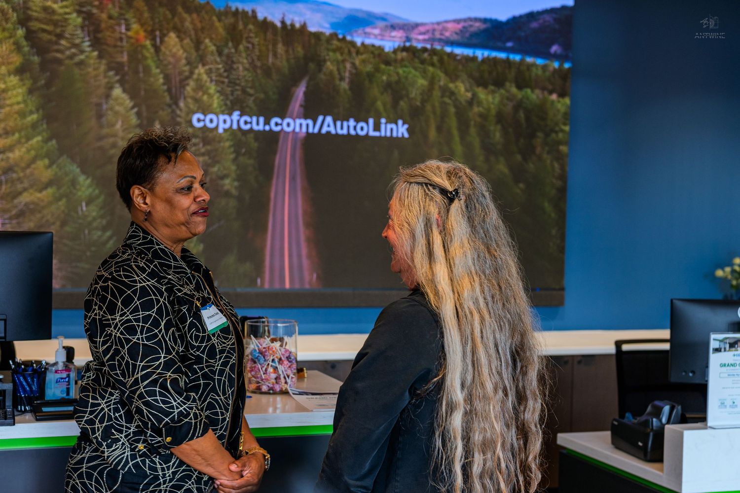 Two women in conversation at COPFCU credit union branch with AutoLink service URL displayed on screen behind them.