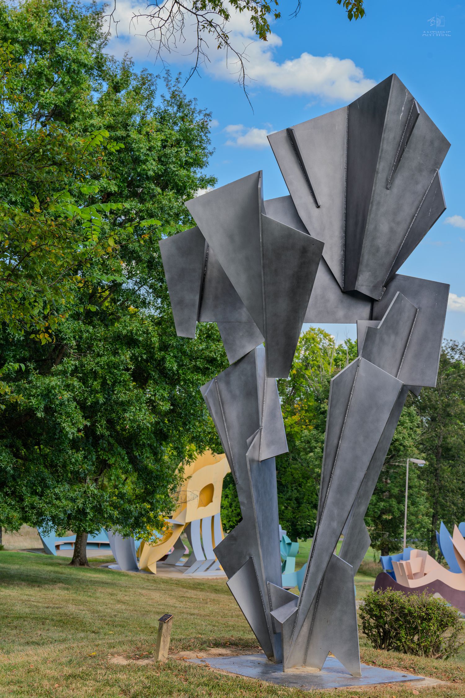 Large abstract metal sculpture with angular geometric shapes displayed outdoors in a park with green trees and blue sky.