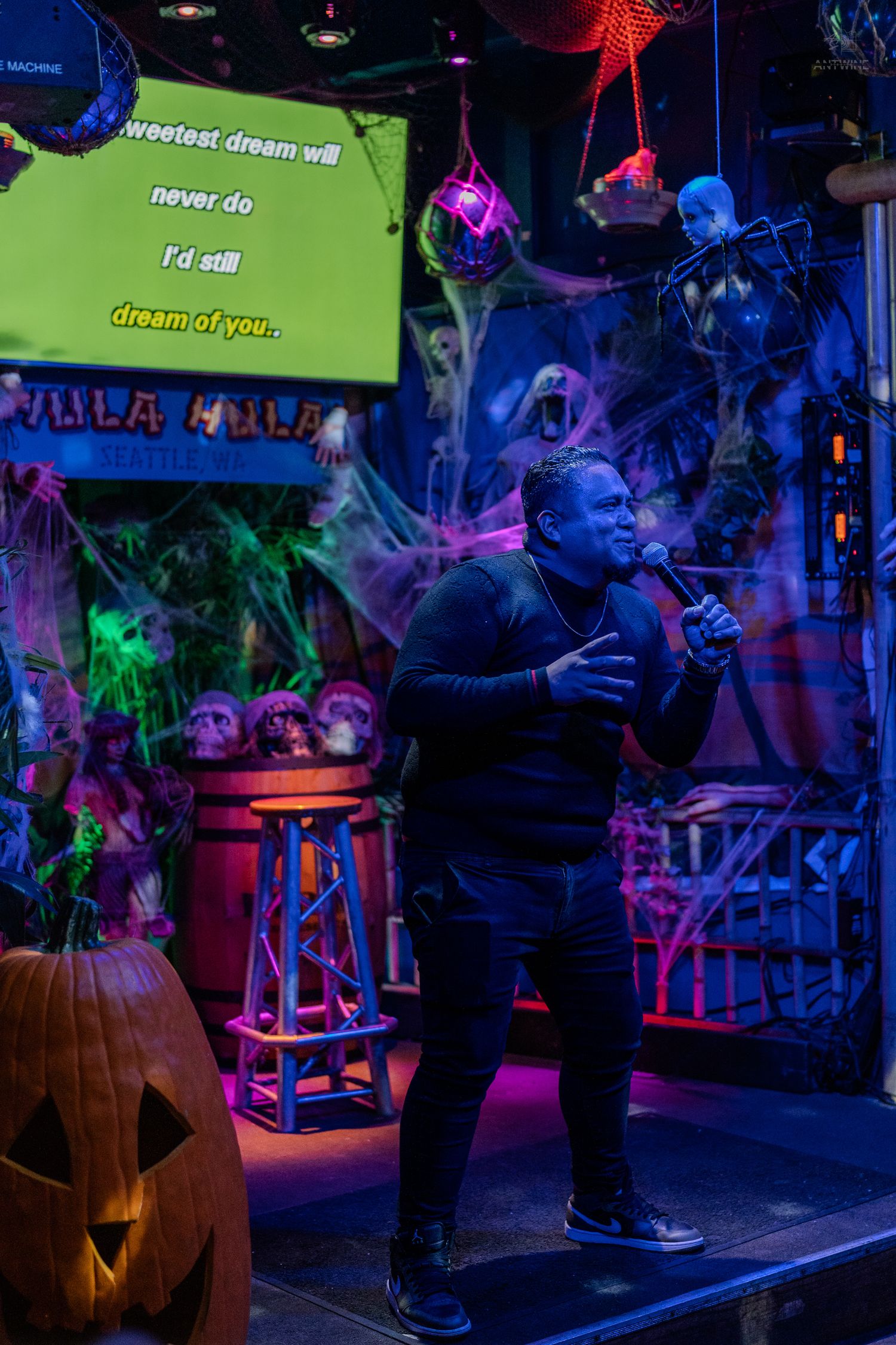 Man in all black singing karaoke on a Halloween-decorated stage at Hula Hula bar in Seattle, WA.