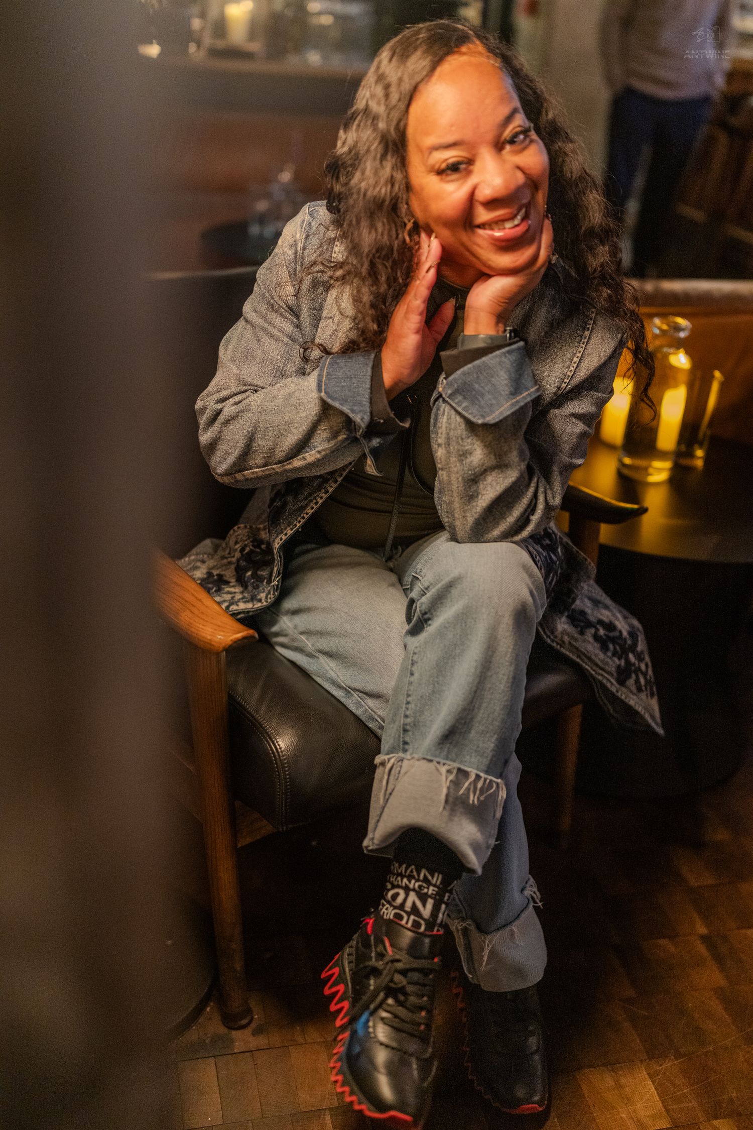 A smiling woman in denim jacket and jeans sits relaxed in a leather chair at a cozy restaurant.