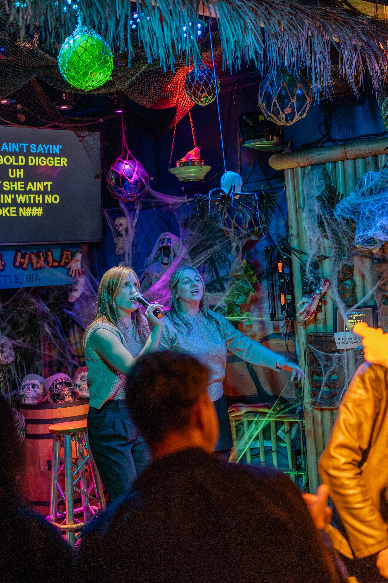 Two women singing karaoke on a colorful tiki bar stage with neon lights and Halloween decorations.