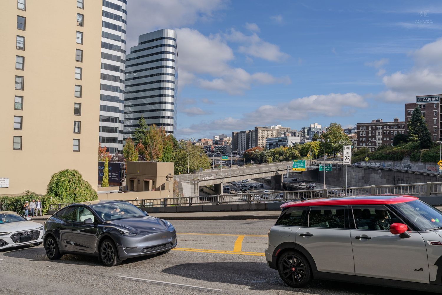 Tesla and Mini Cooper cars driving on a Seattle urban highway with residential buildings and blue sky.