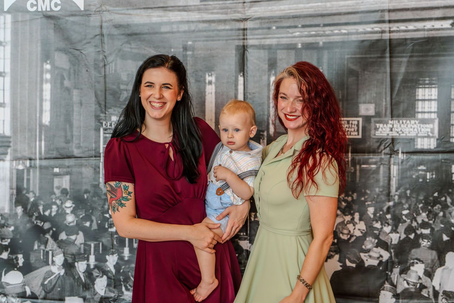 Two women and a baby posing indoors in front of a vintage black and white photograph backdrop.