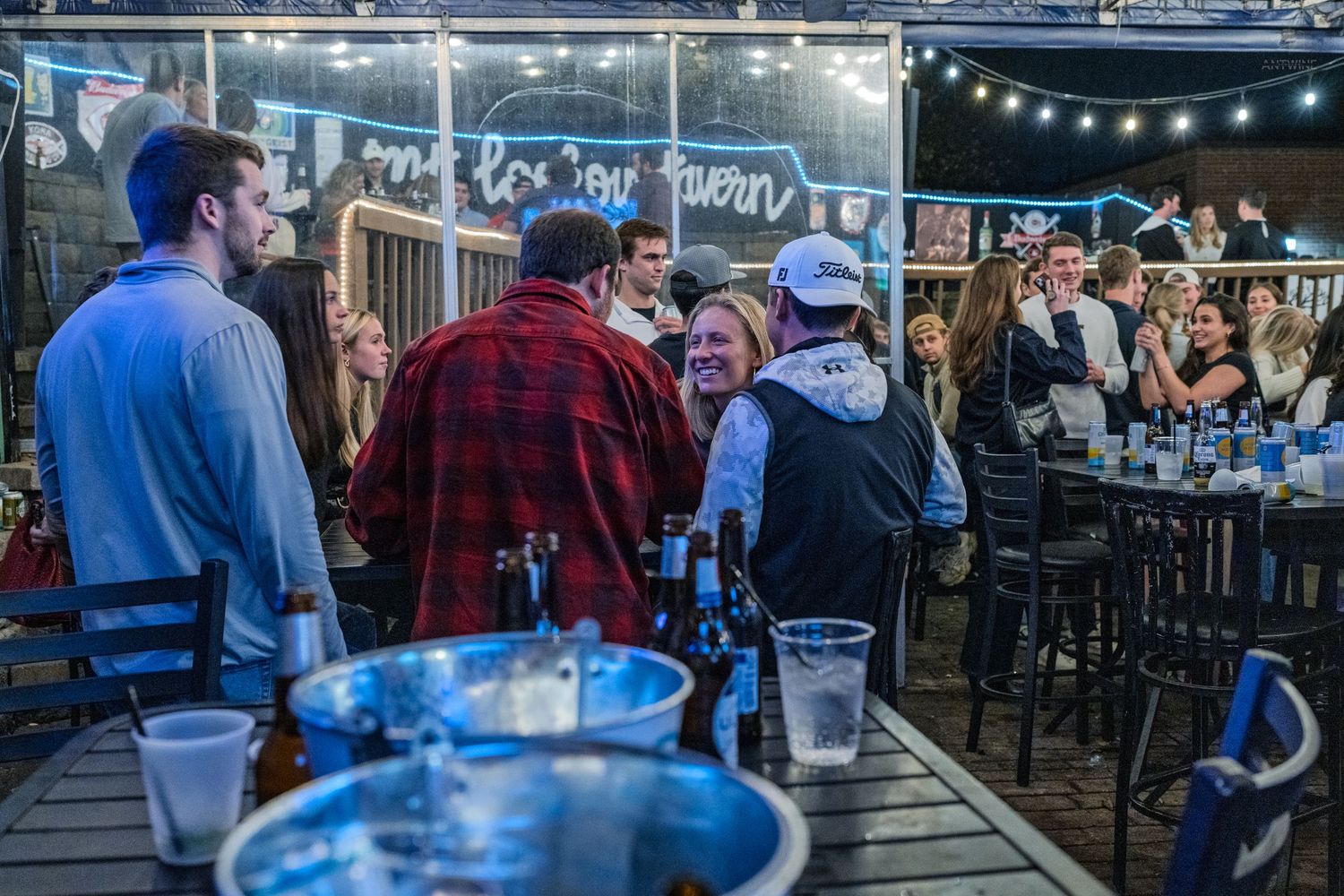 Group of friends socializing and laughing at an outdoor bar patio with string lights at night.