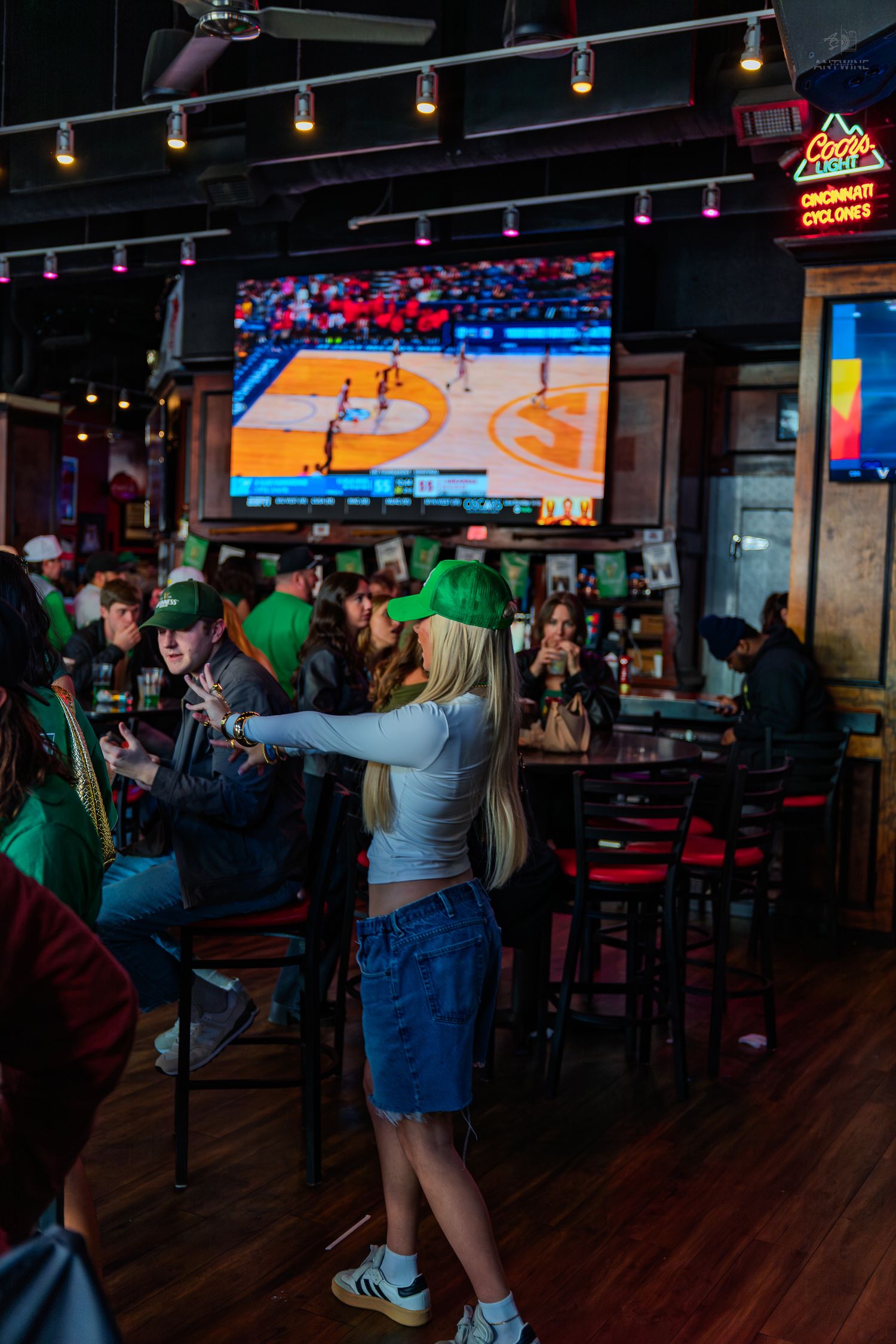 Blonde woman in green hat and denim skirt standing in a busy sports bar watching basketball on a large screen TV.