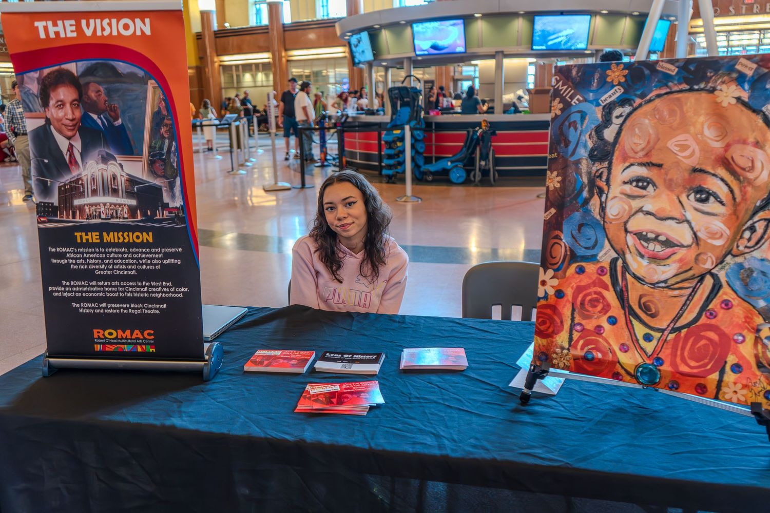 Woman sitting at a booth with colorful artwork and mission display banner at an indoor event.