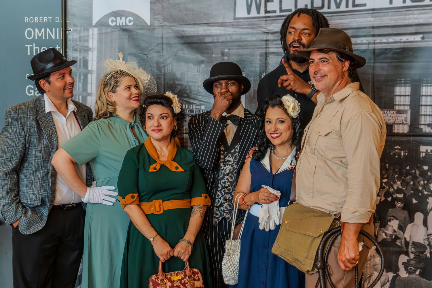Group of seven actors in 1940s vintage costumes posing together at a theater event near a Welcome sign.
