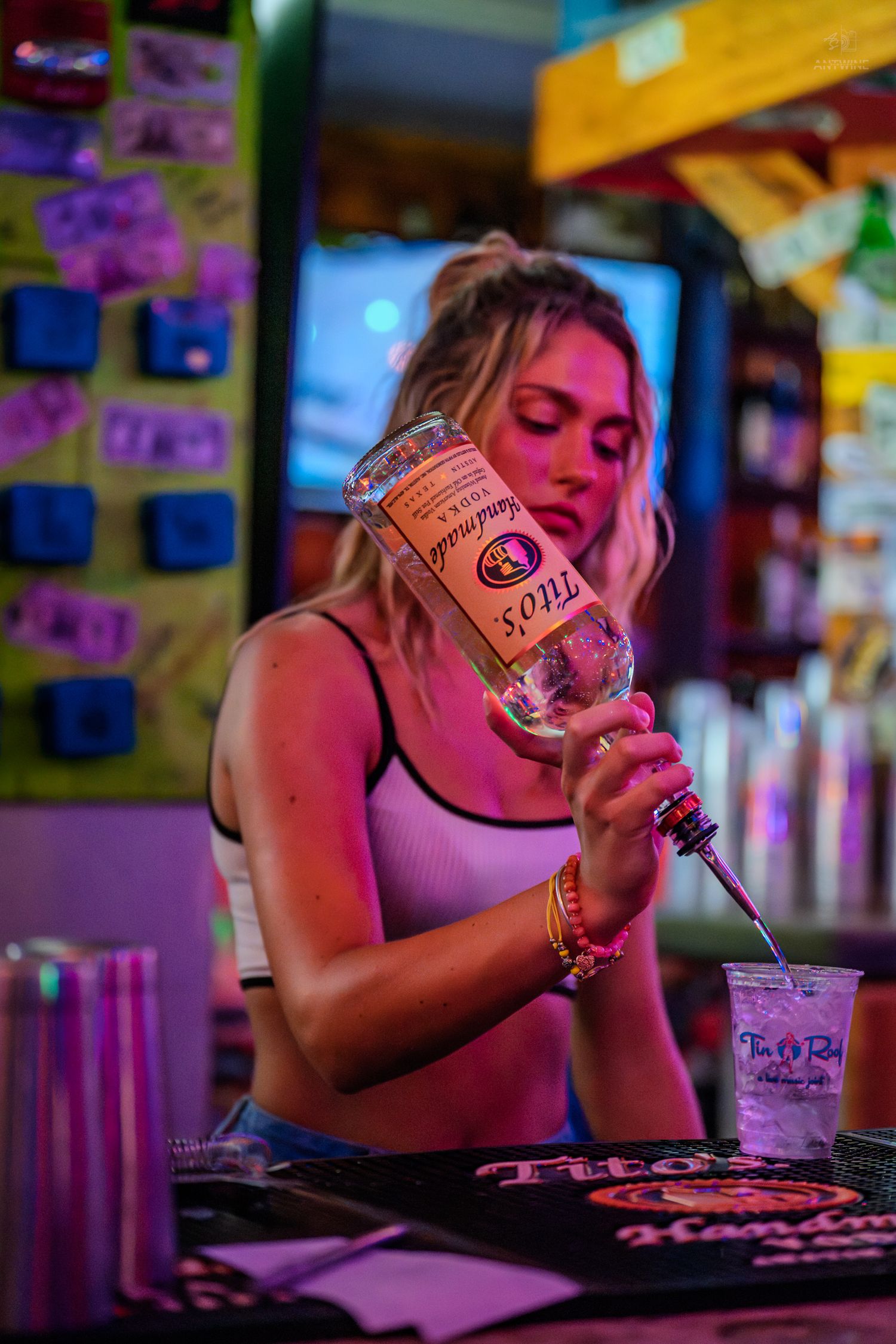 Blonde bartender pouring Titos Handmade Vodka into a cup at a vibrant neon-lit bar.