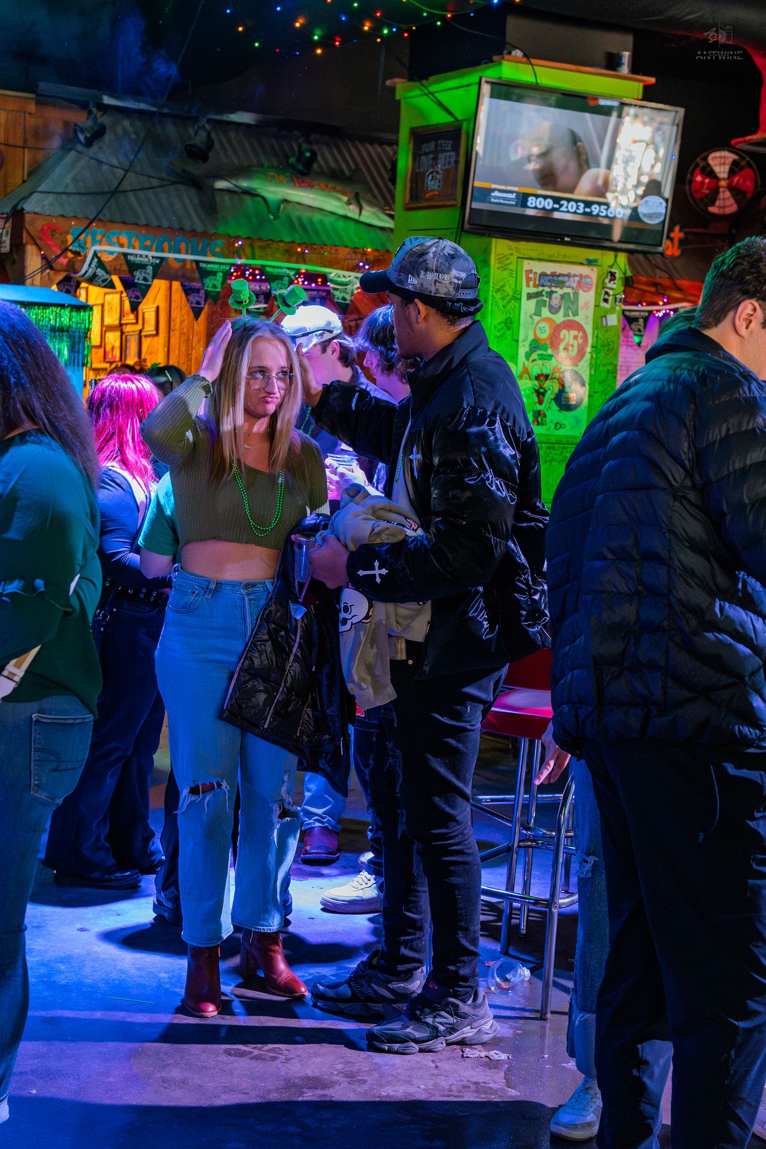 People socializing in a vibrant neon-lit bar with green decor, a woman in crop top and jeans stands out.