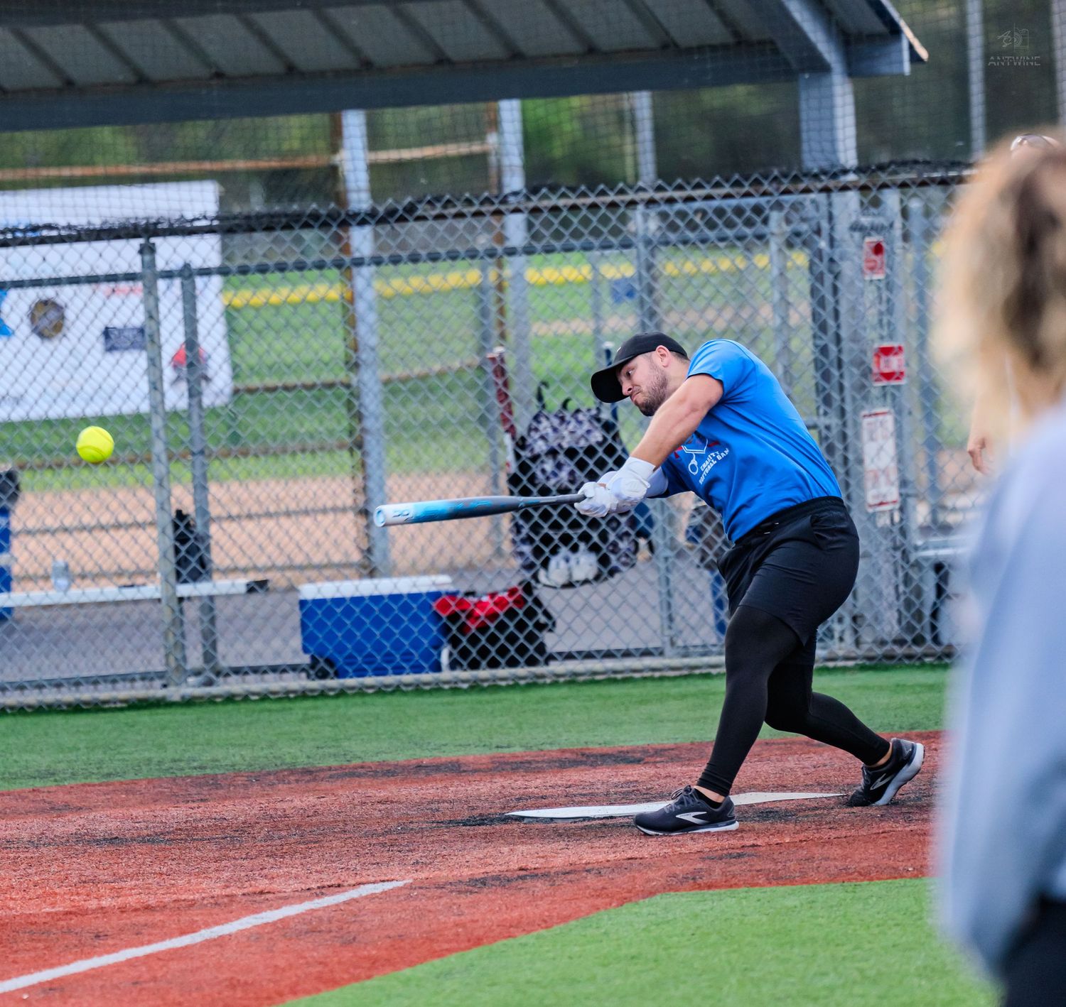 A softball player in a blue jersey swings a bat at an incoming yellow ball on a red clay diamond.