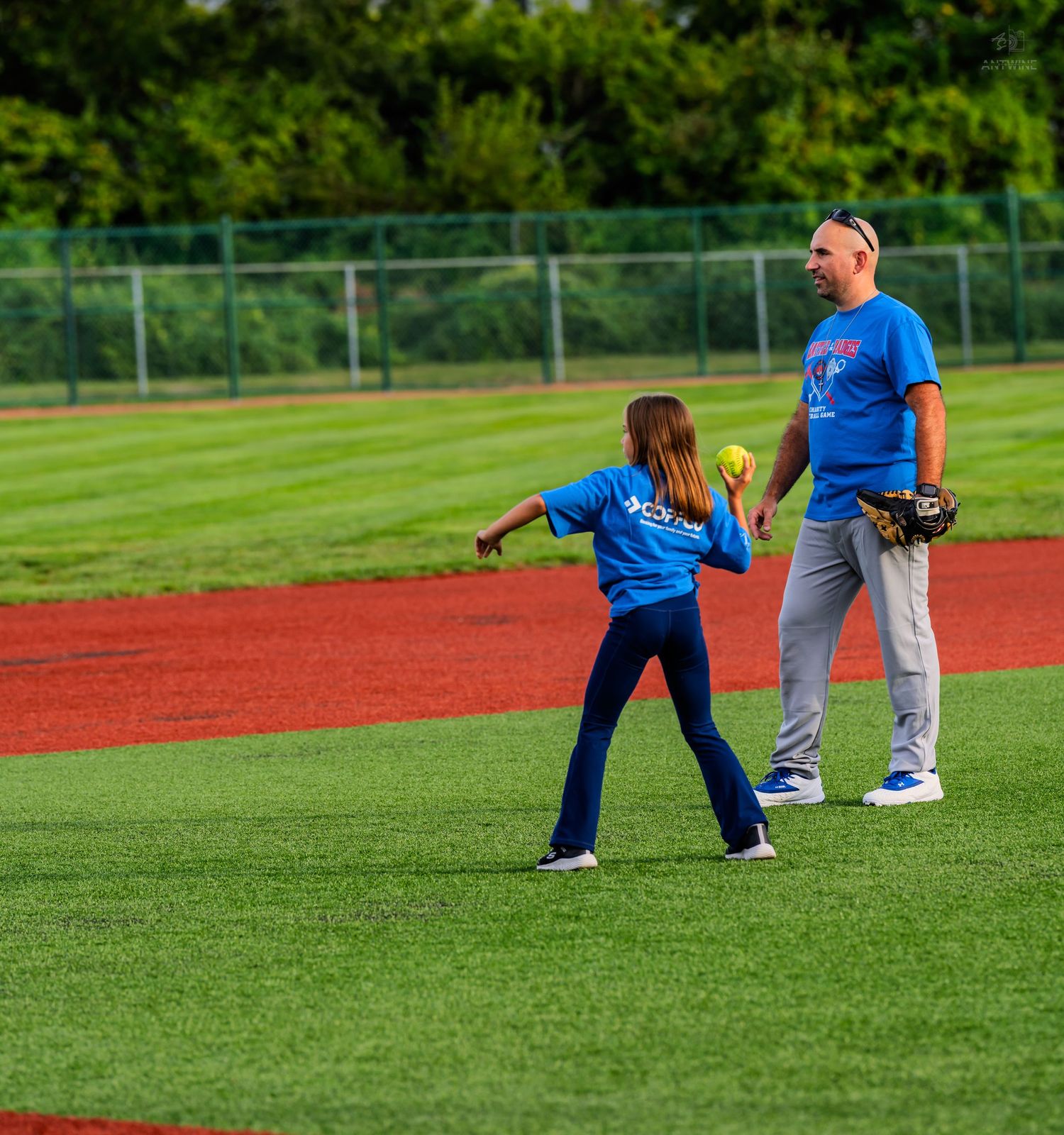 Young girl in blue shirt throwing a yellow softball while adult coach watches on green field.