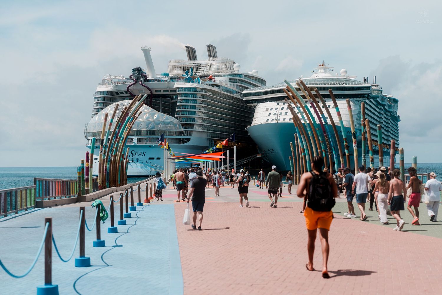 Tourists walking along a colorful cruise ship terminal boardwalk with large Royal Caribbean ships docked in the background.