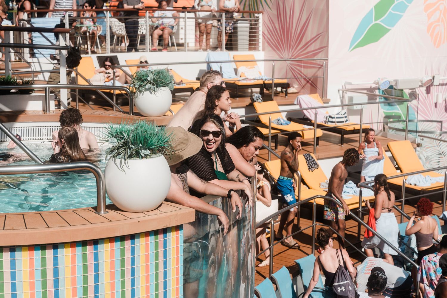 Guests relax and socialize on a colorful cruise ship pool deck with lounge chairs and tropical decor.