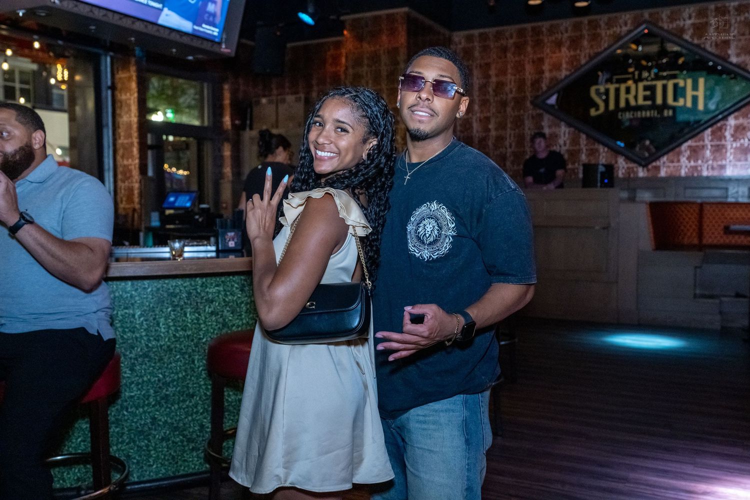 A smiling couple poses together at a bar, with a Stretch sign visible in the background.