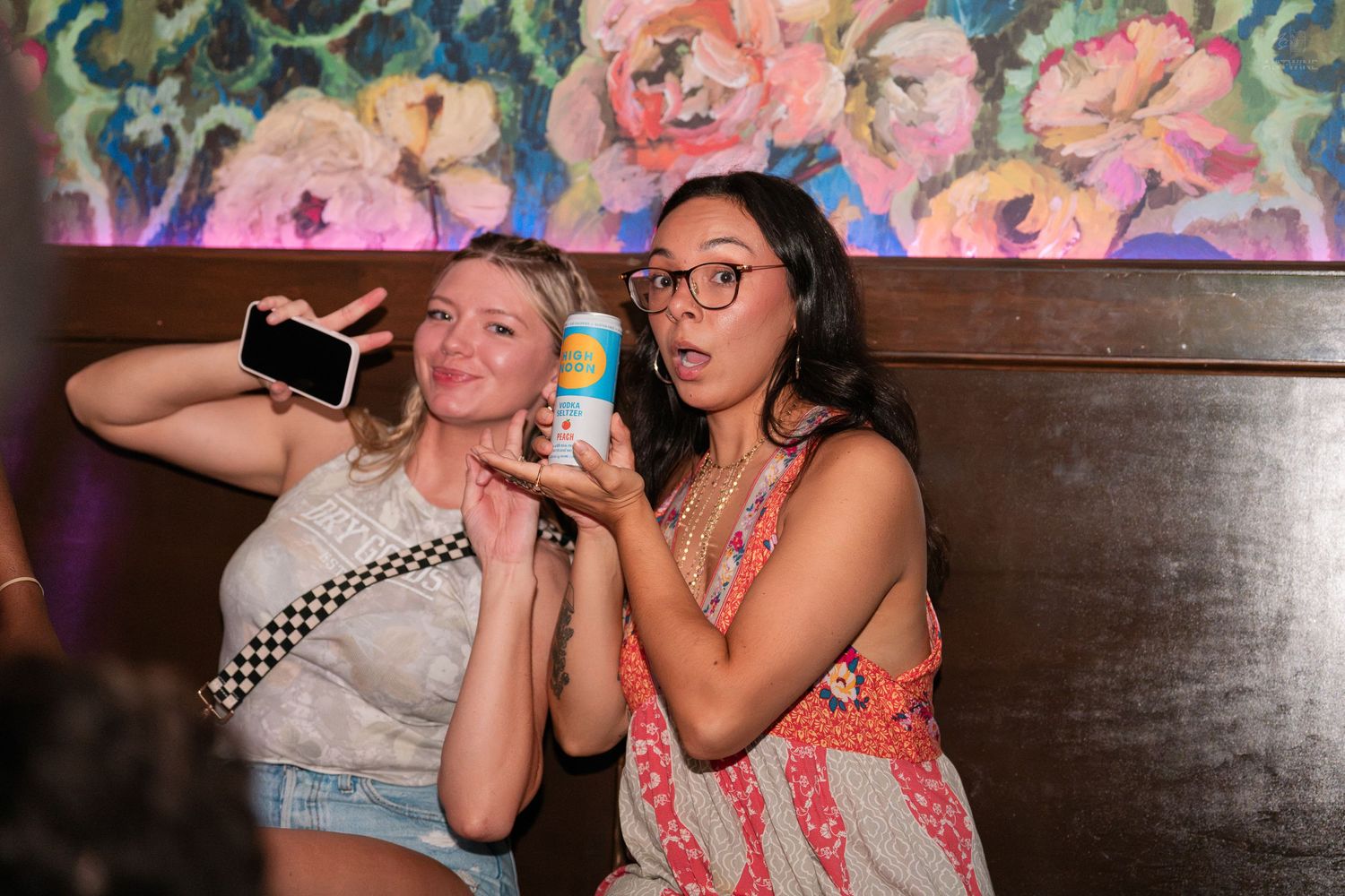 Two women posing playfully at a bar, one holding a phone and a canned drink.