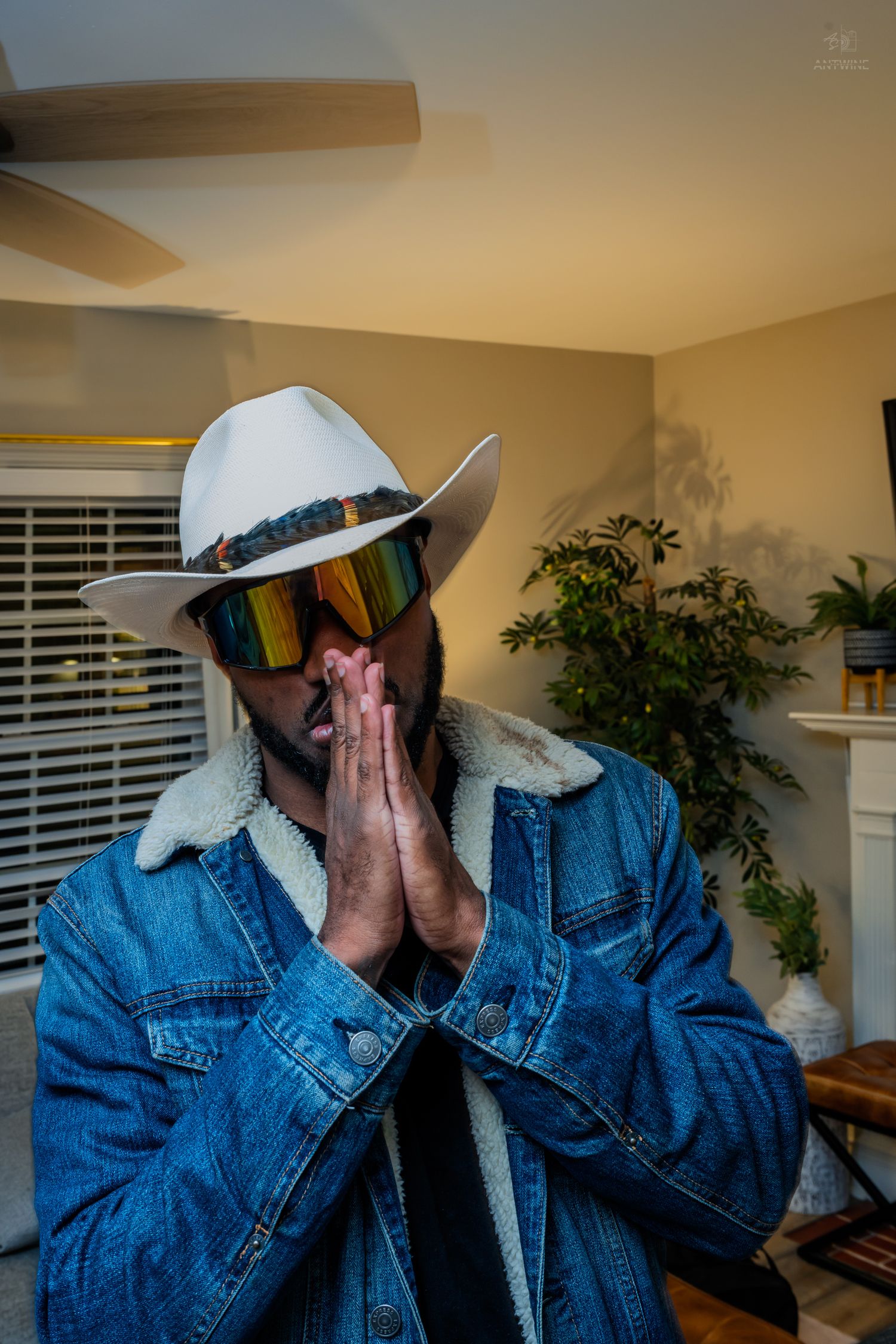 Man in white cowboy hat and rainbow mirrored sunglasses wearing denim jacket with hands in prayer pose.