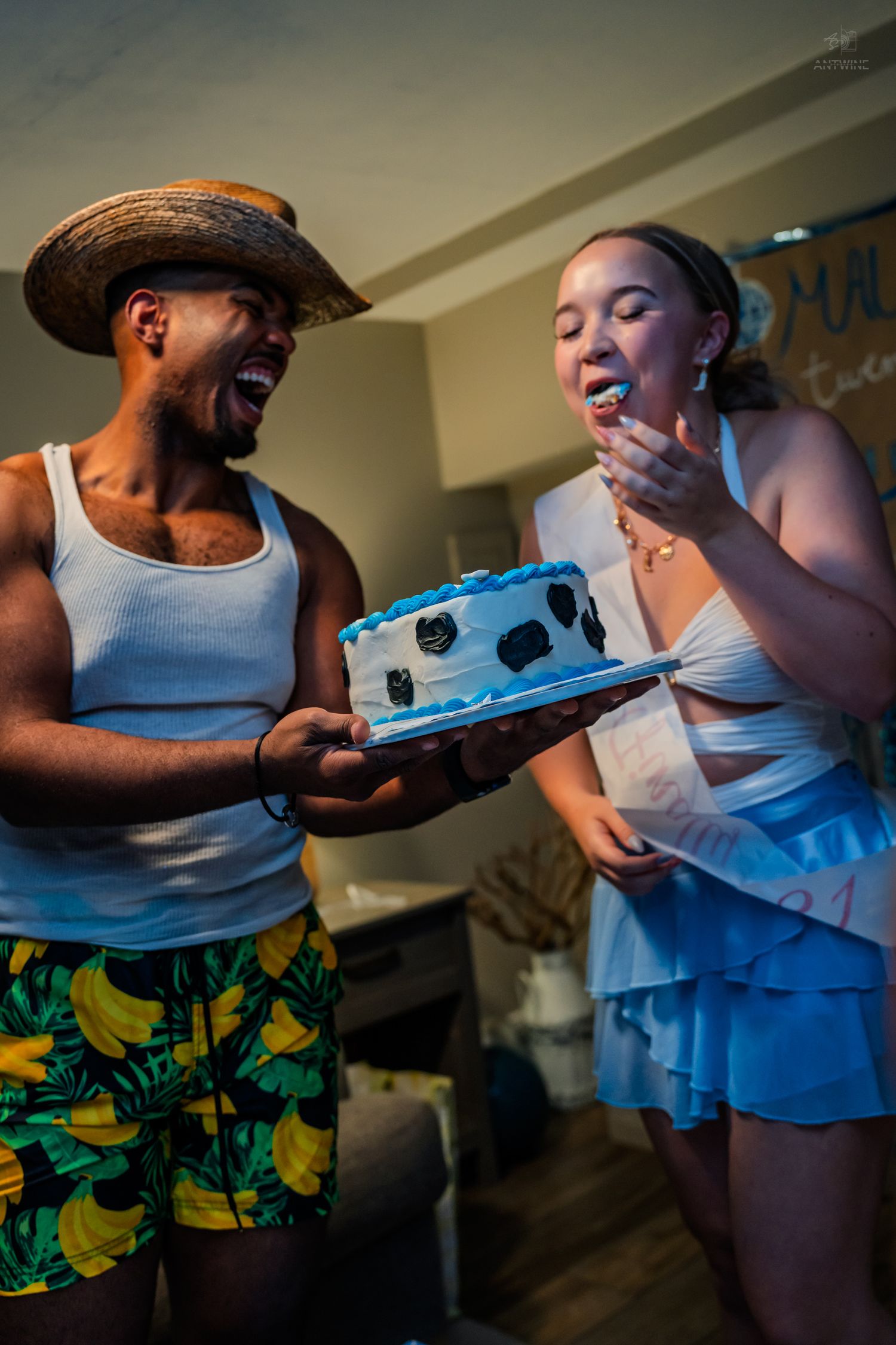 Man in straw hat laughs as woman eats frosting from a blue decorated birthday cake indoors.