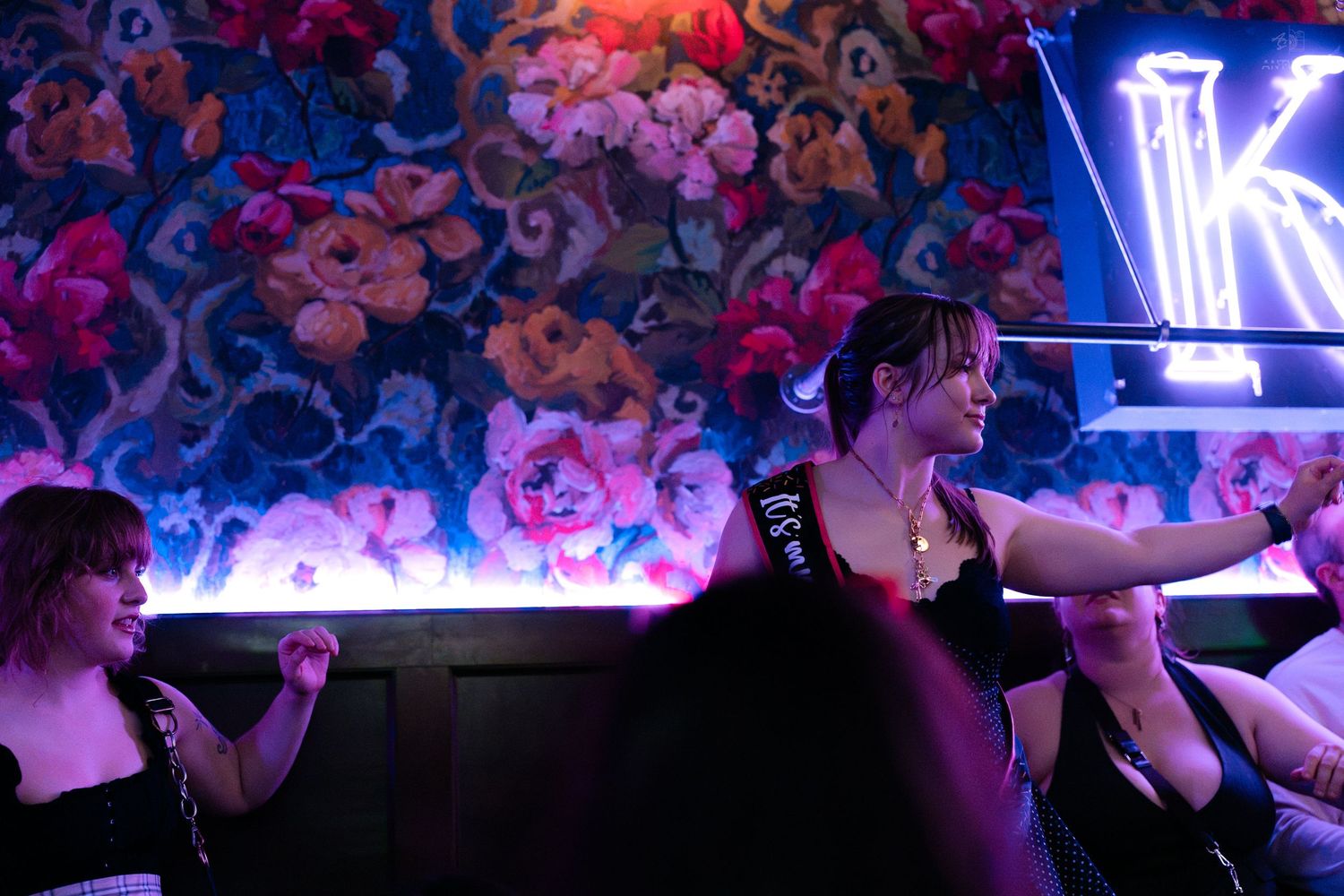 Women dancing at a lively nightclub with neon lights and vibrant floral wallpaper decor.
