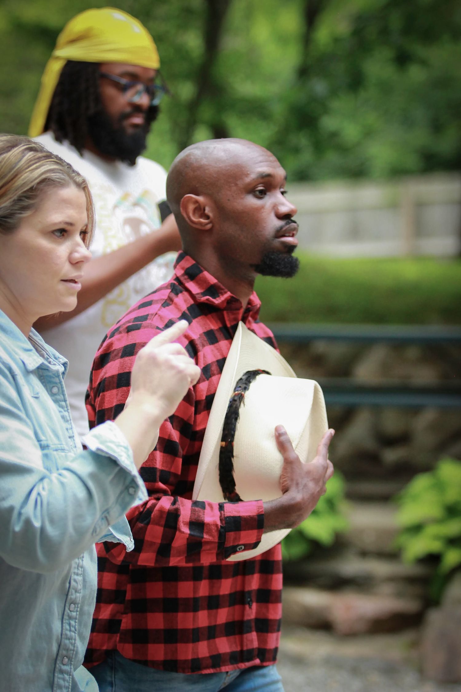 Three people stand outdoors in a wooded area, with a man in a red plaid shirt holding a straw hat.
