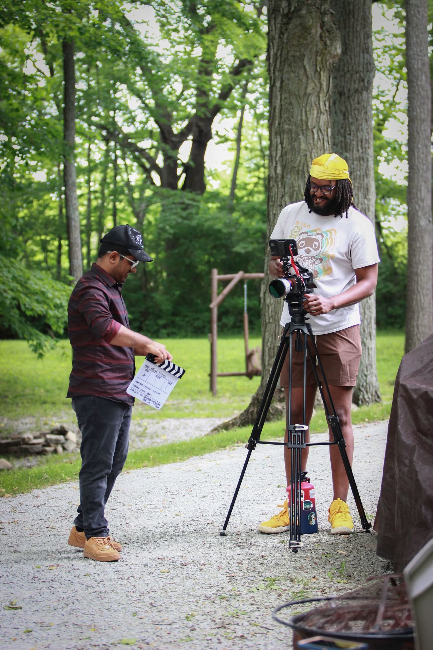 Two filmmakers work outdoors on a wooded path, one holding a clapperboard while the other operates a camera on a tripod.