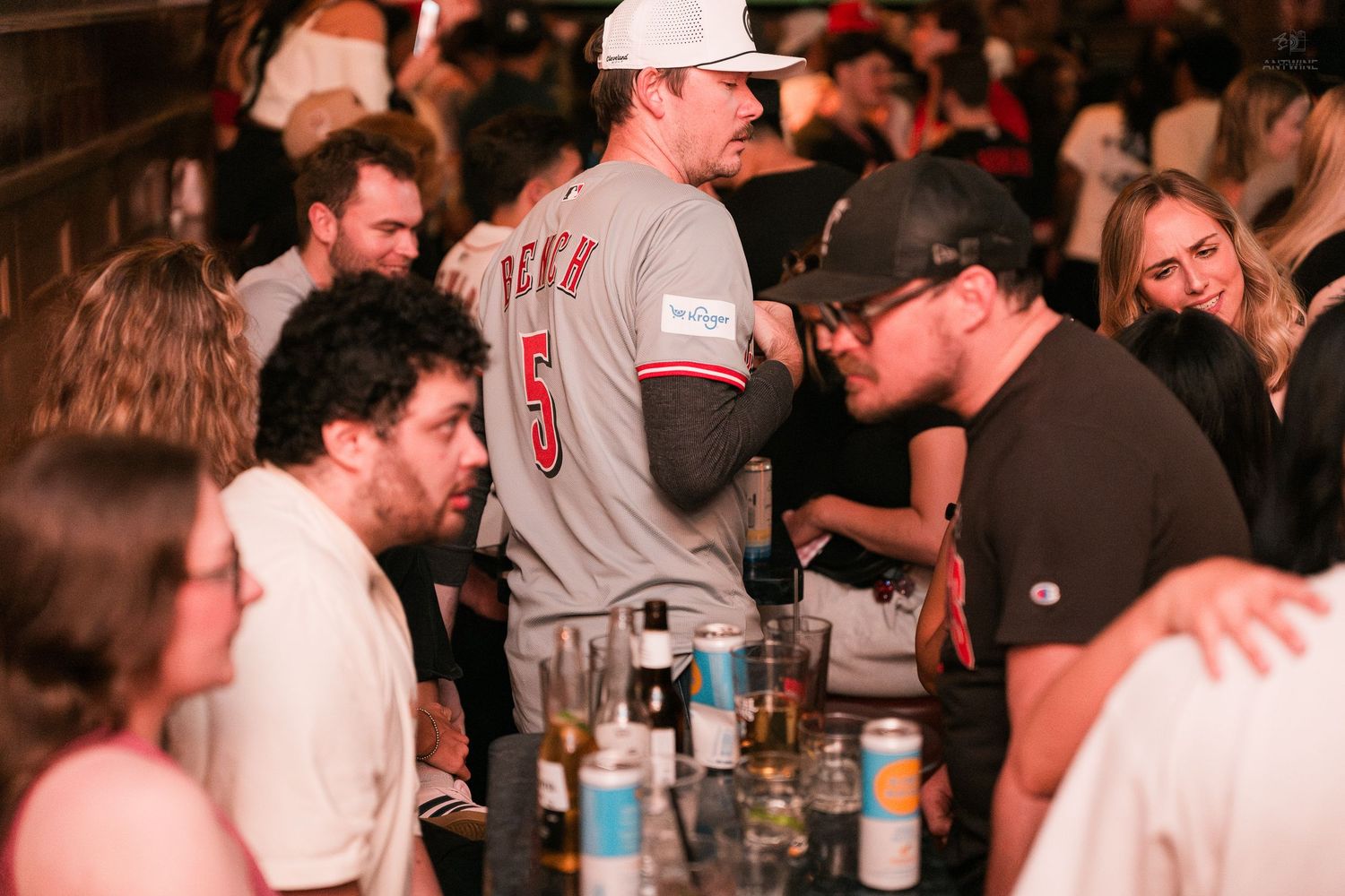 Man in Cincinnati Reds jersey number 5 socializing at a crowded bar with friends and drinks on the table.