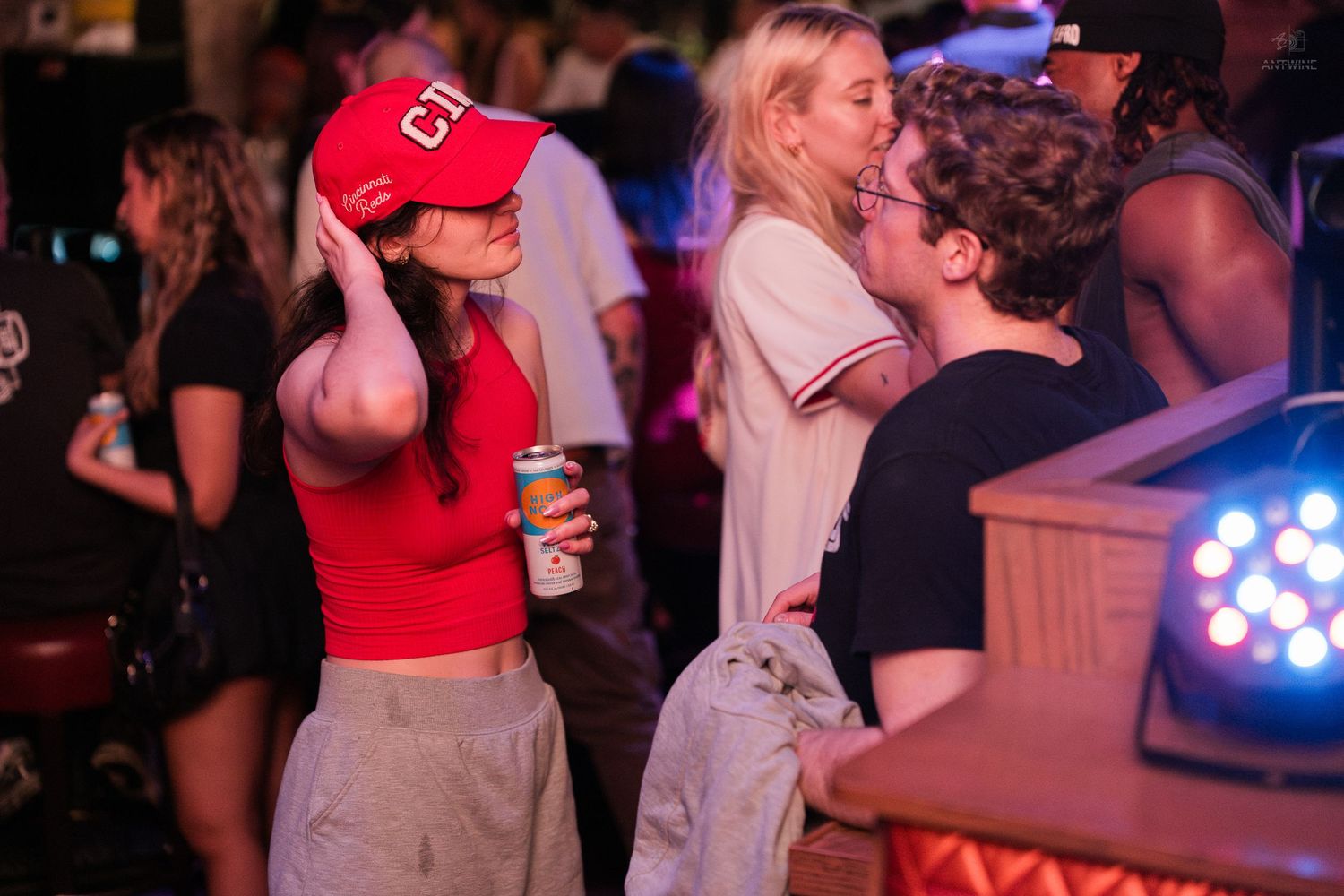 Young woman in red Cincinnati Reds cap holds drink while socializing at crowded sports bar.