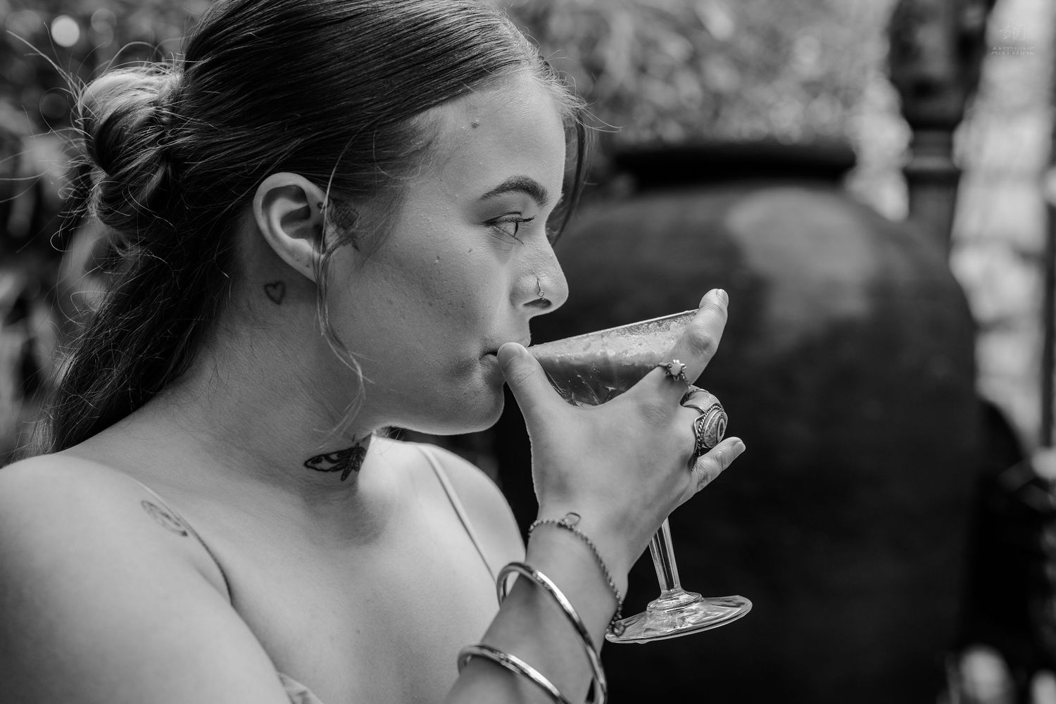 Black and white photo of woman drinking from martini glass with gold jewelry outdoors.