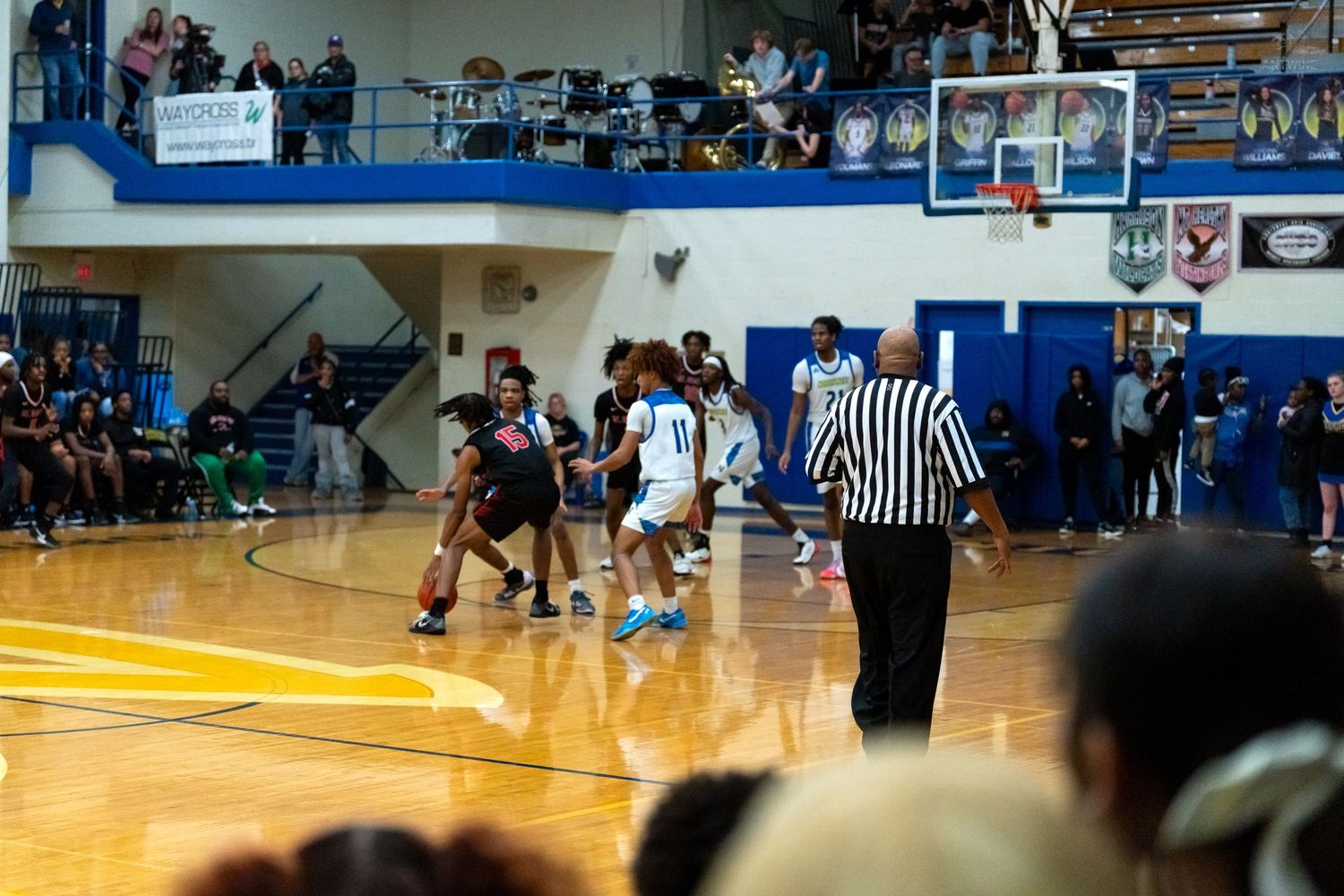 High school basketball game in progress with players, referee, and crowded gymnasium bleachers in background.