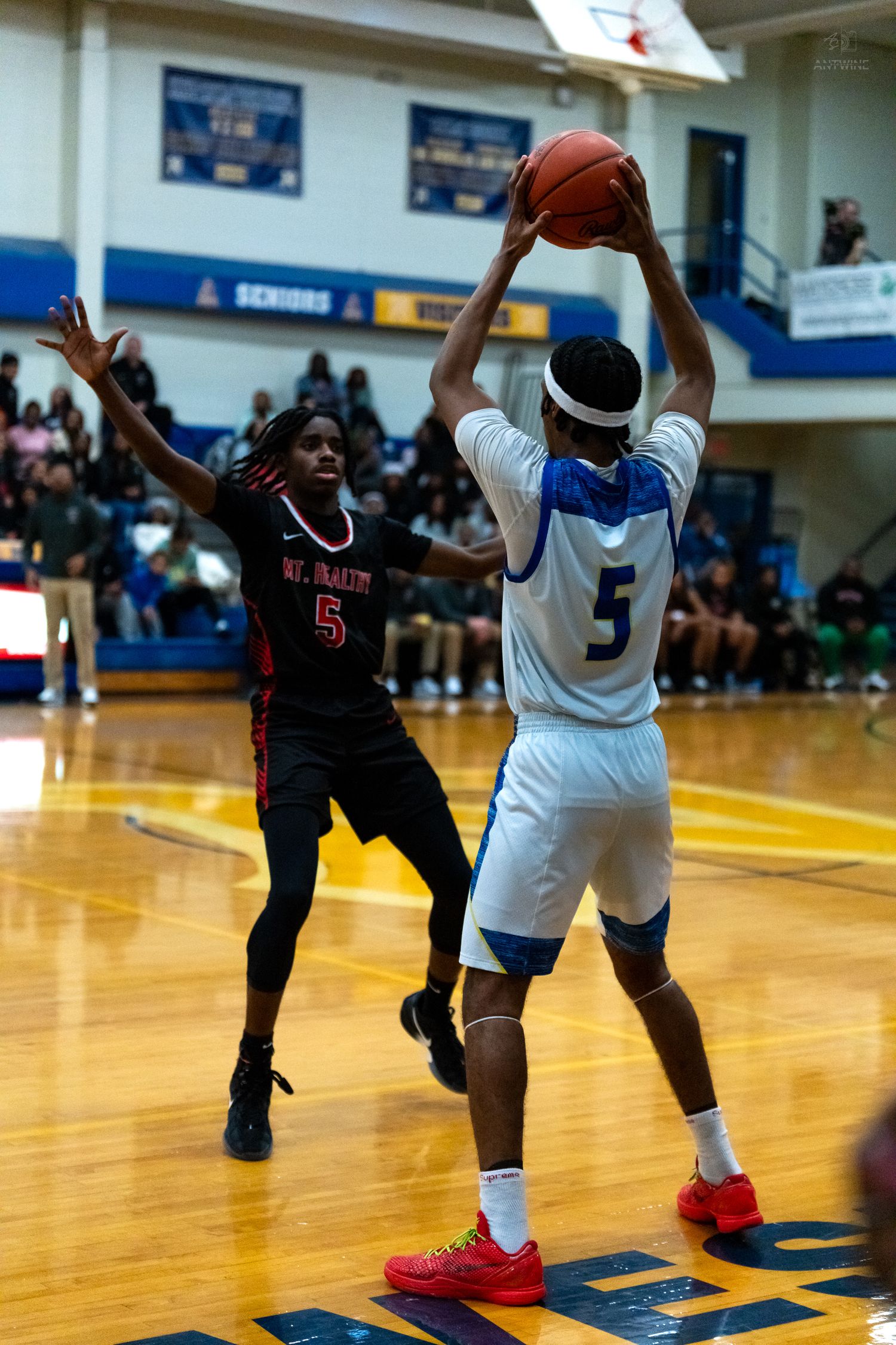 Player number 5 in white uniform rises to shoot over defender number 5 in black during high school basketball game.