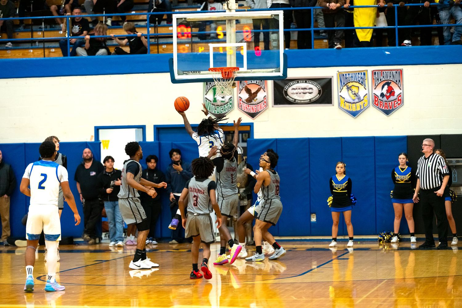 Basketball player attempts layup during high school game in blue and white gym with championship banners.