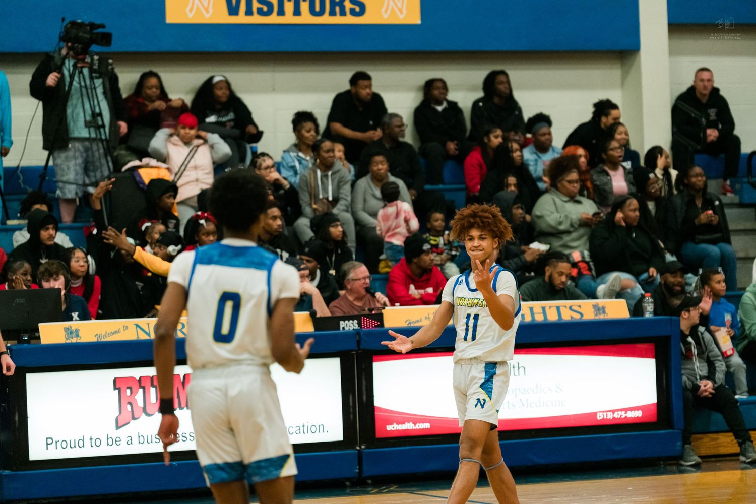 Two basketball players in blue and white uniforms celebrate with a high-five in front of the visitors scoreboard.