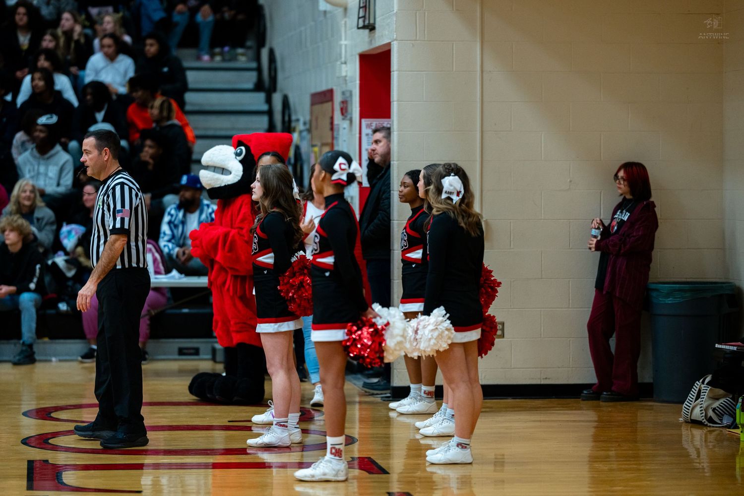 Cheerleaders in black uniforms line sideline beside referee during packed high school basketball gymnasium game.