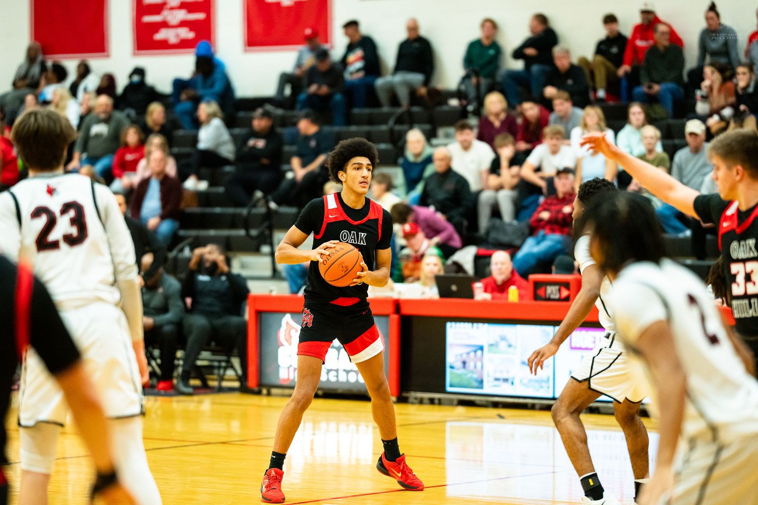 A basketball player in red holds the ball while surrounded by defenders in a crowded gym game.