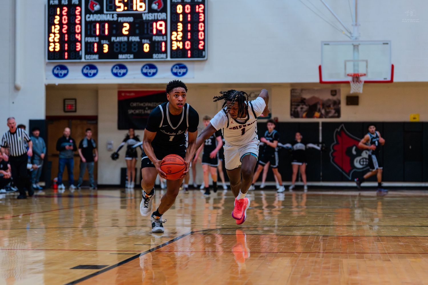 Two basketball players race down the court during a high school basketball game with scoreboard visible.
