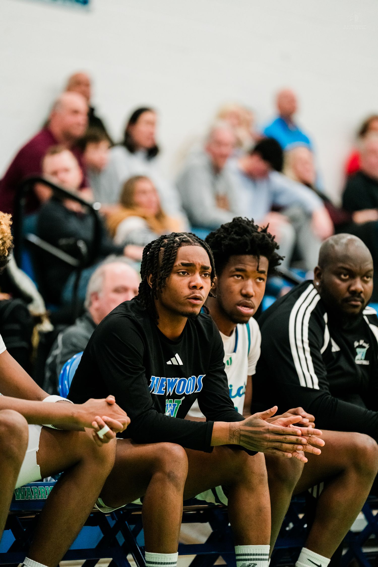 Three players in black Lakewood warmups sit focused on the bench watching a high school basketball game.