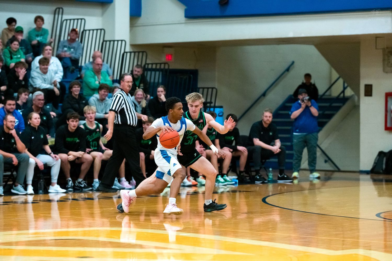 A blue-uniformed player dribbles upcourt while a green-jersey defender applies pressure in a packed gymnasium.