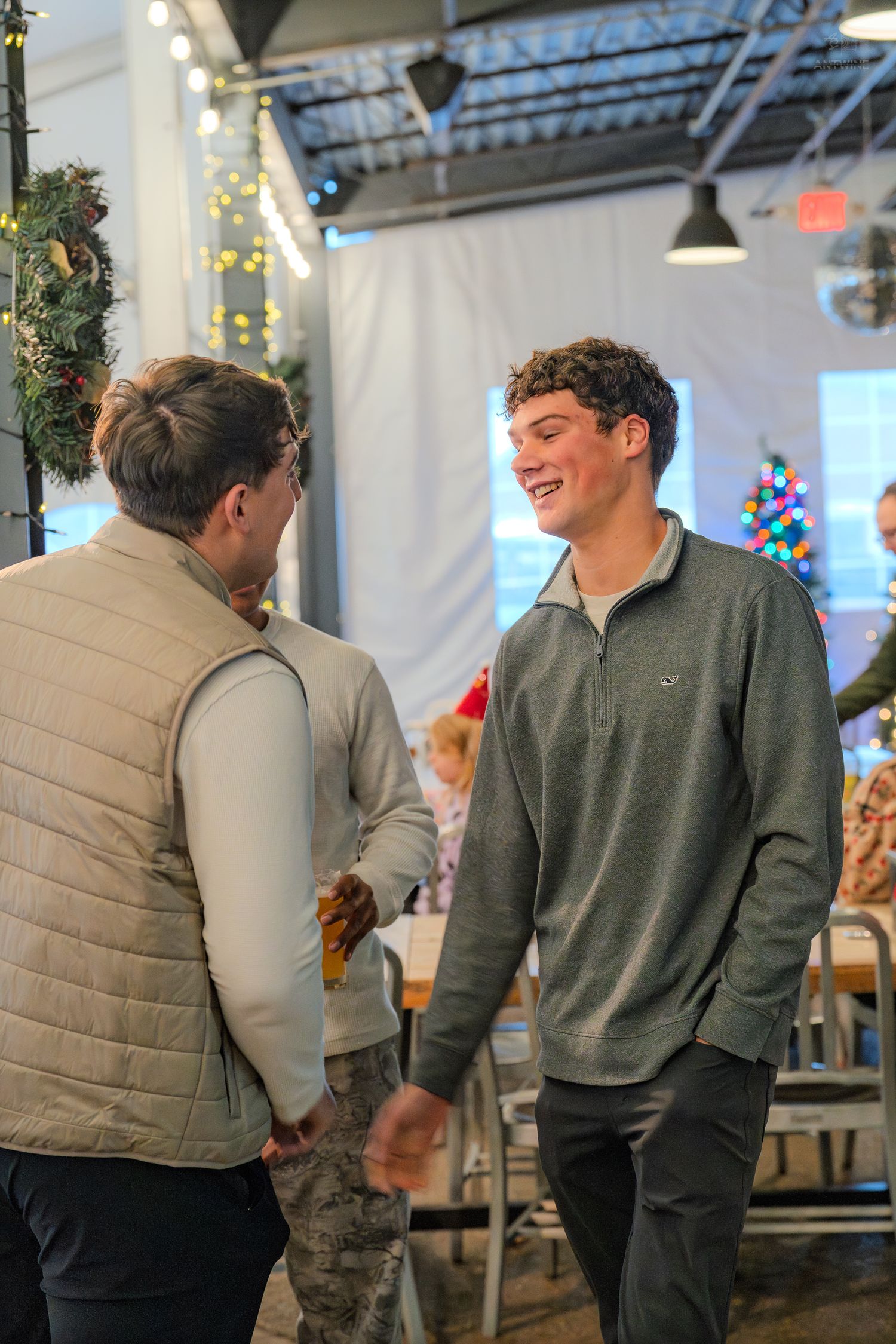 A smiling young man in a gray pullover shakes hands with another man at a festive holiday party indoors.