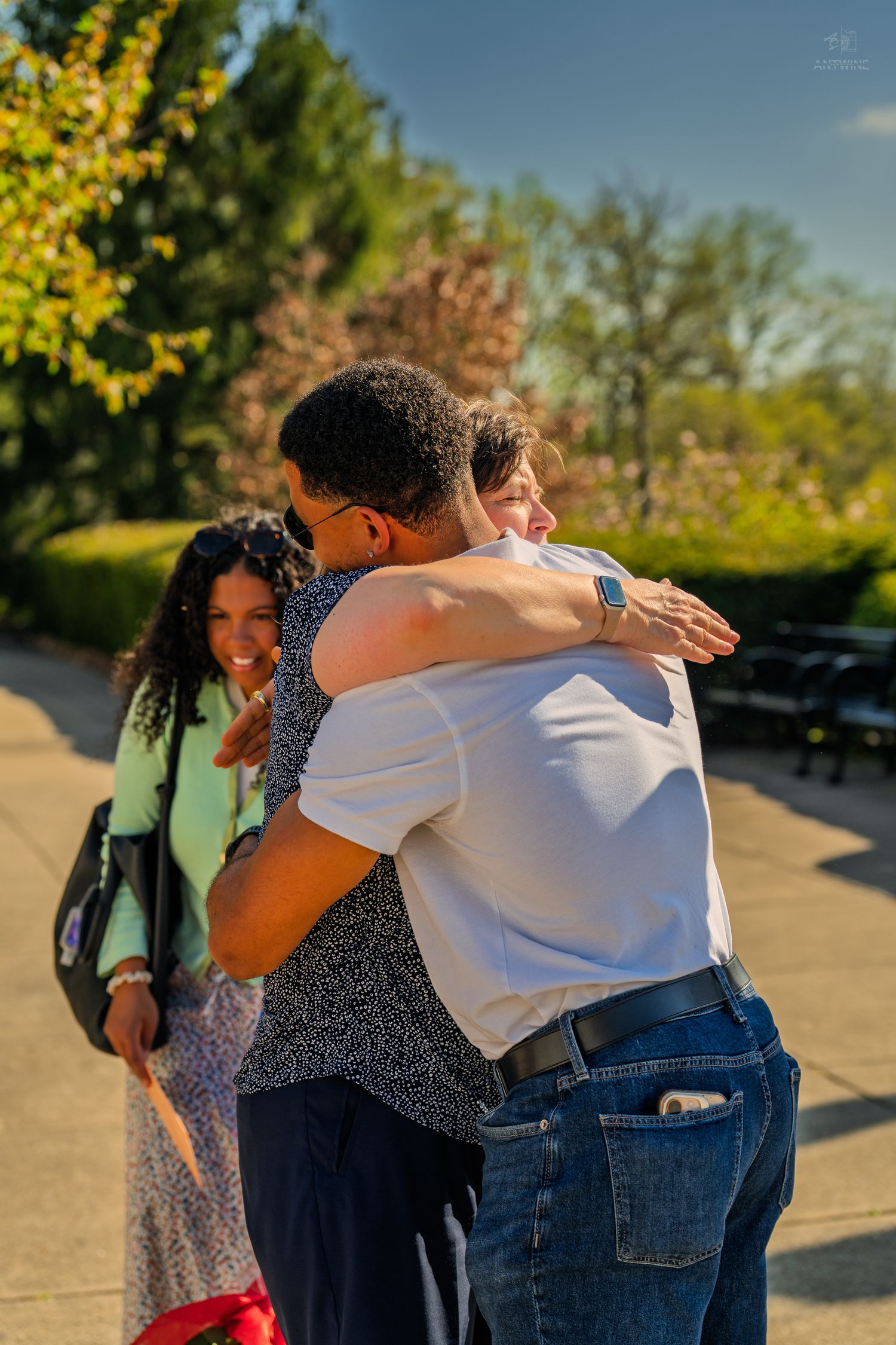 Two people hug warmly outdoors in sunlight while a smiling woman in green jacket looks on nearby.